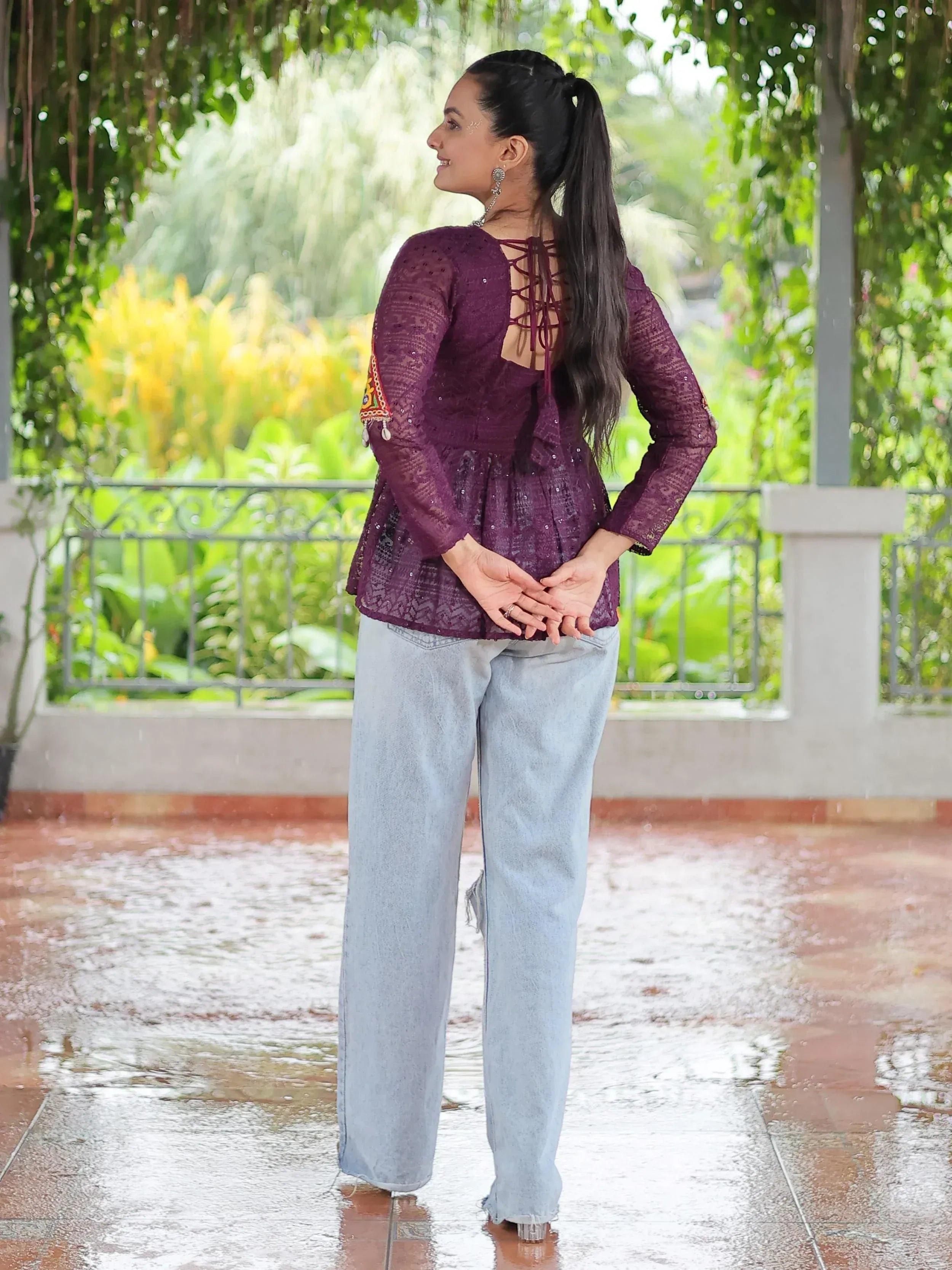 Woman in purple ethnic lace top with back tie detail, paired with light denim jeans, standing outdoors in rain