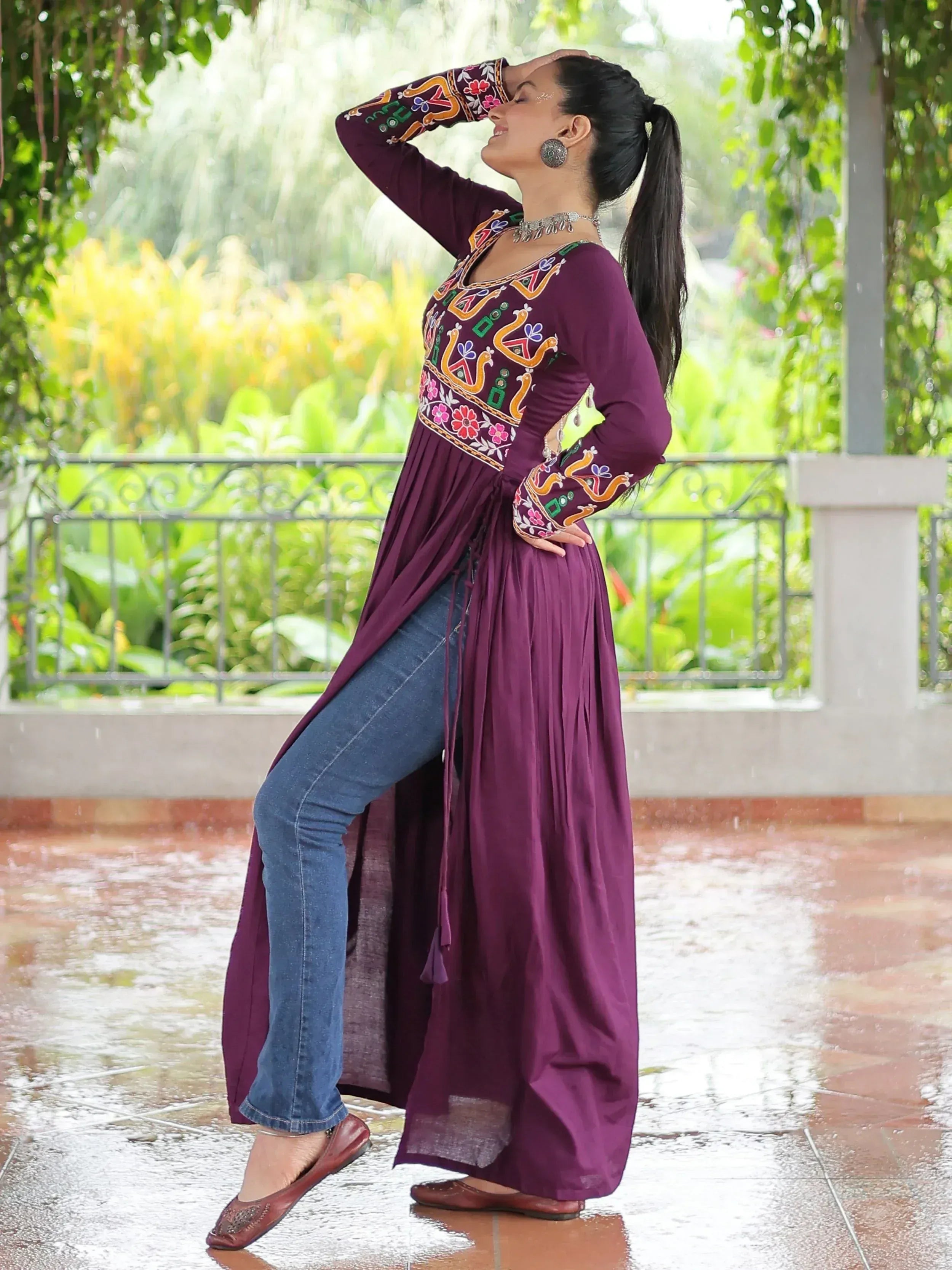 Woman in purple embroidered ethnic kurti with jeans, standing in rain under greenery