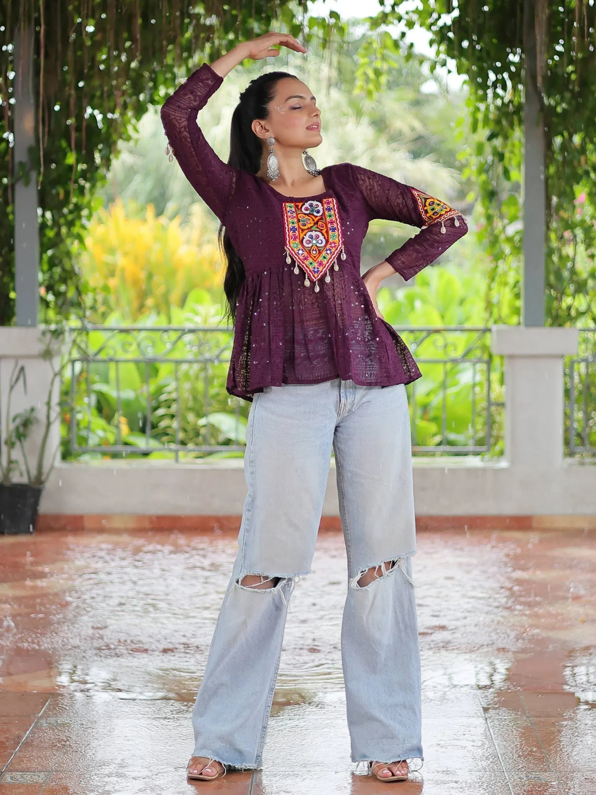 Woman in purple embroidered ethnic top with ripped jeans, dancing in rain under greenery