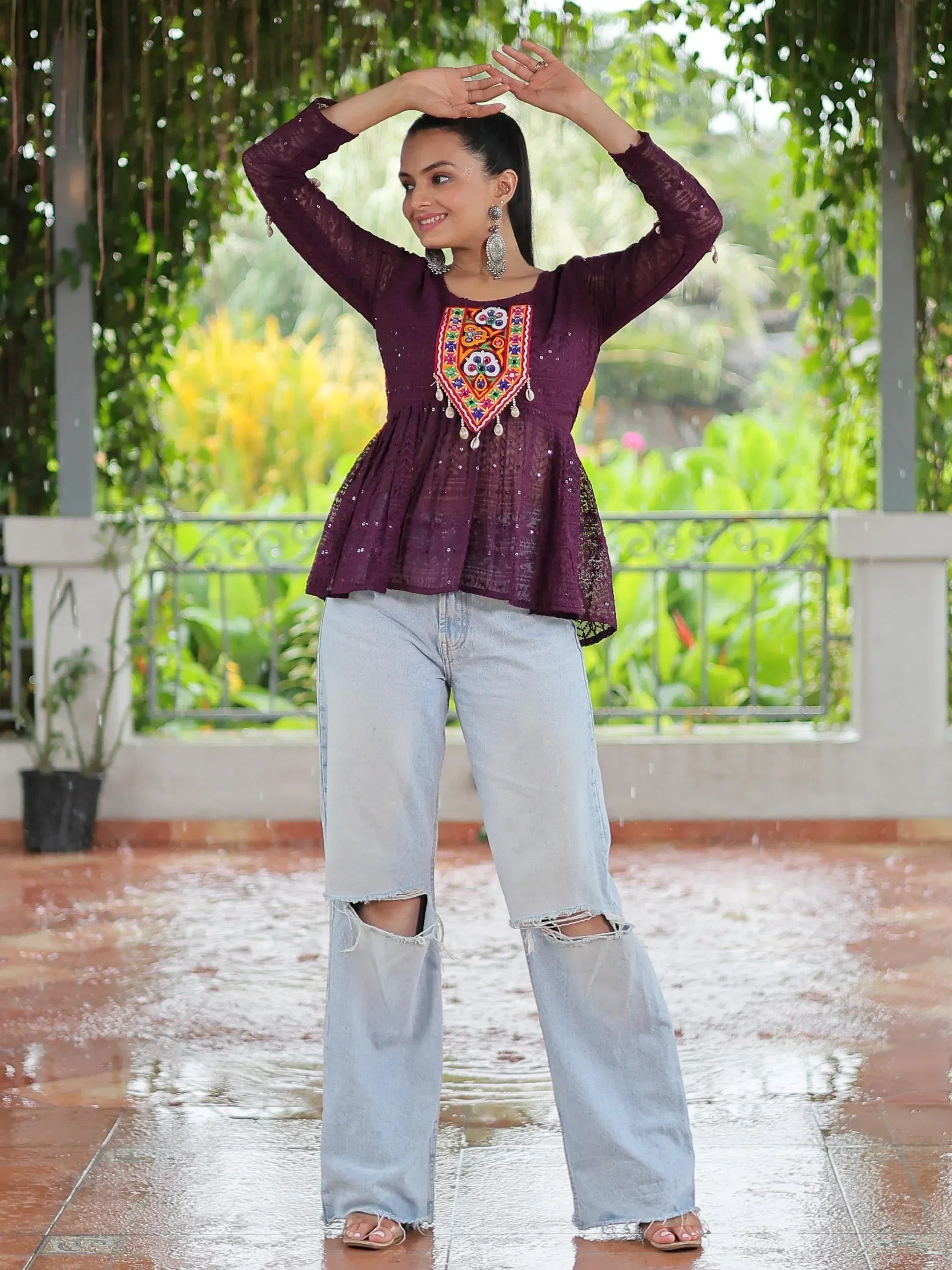 Woman in purple embroidered ethnic top with ripped jeans, dancing in rain under greenery