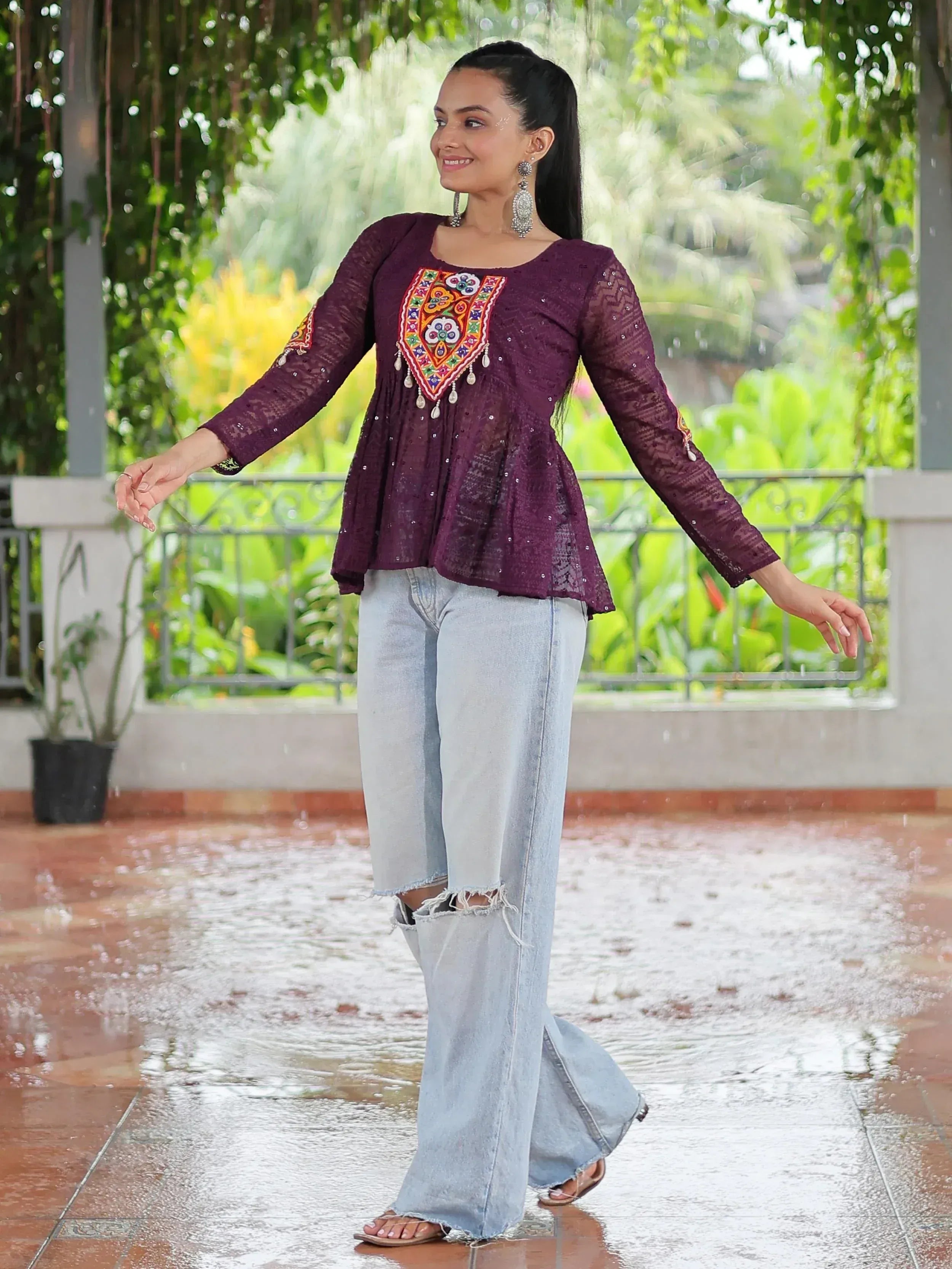 Woman in purple embroidered ethnic top with flared jeans, standing in rain under greenery