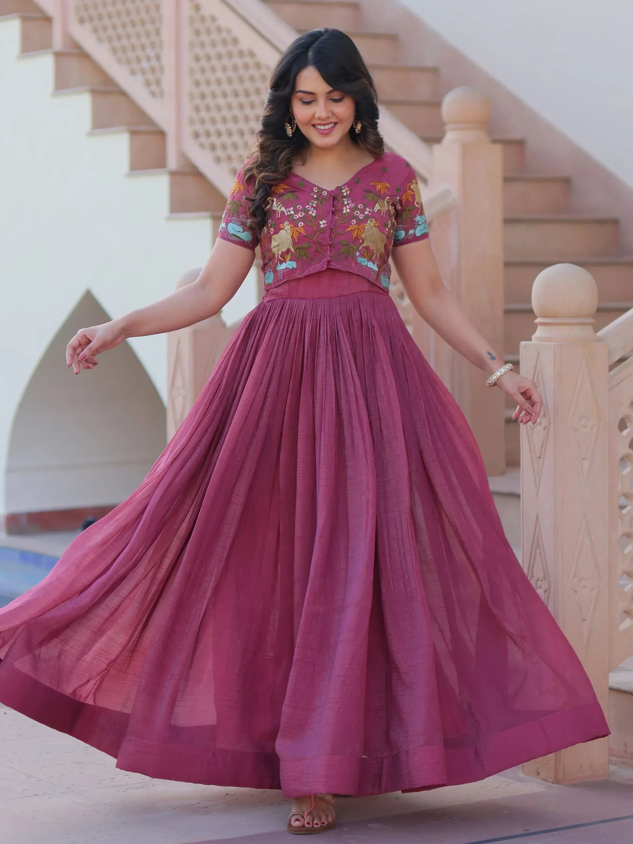 Woman in maroon embroidered ethnic gown with floral and animal motifs, twirling near staircase