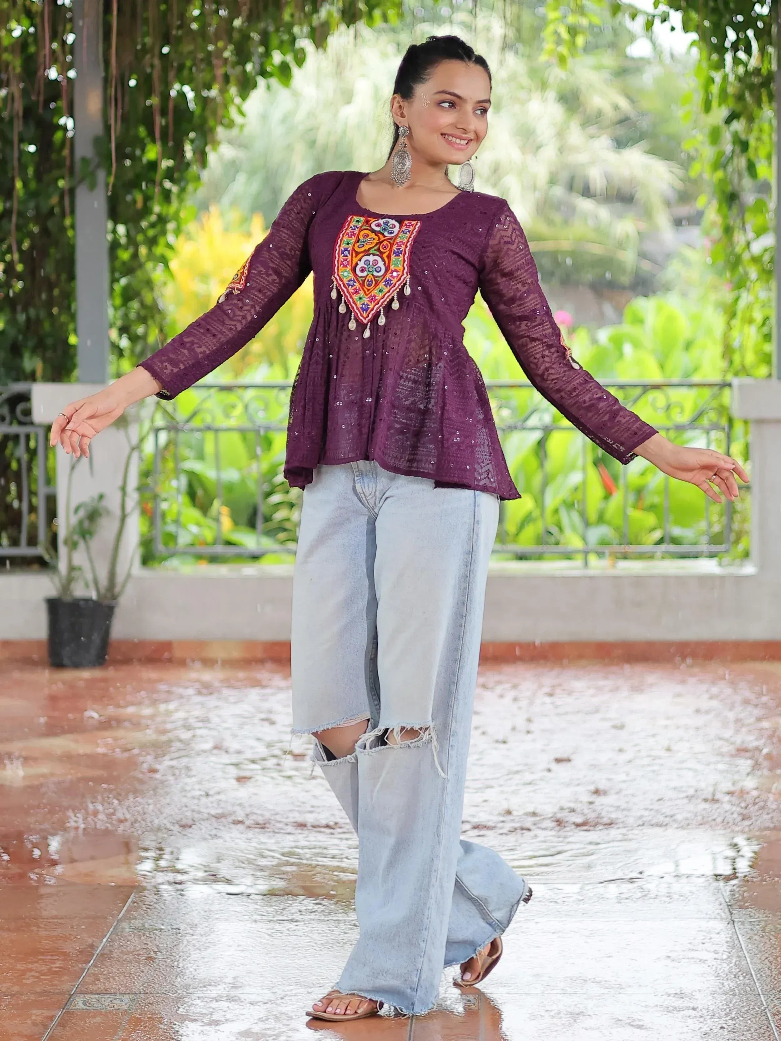 Woman in purple embroidered ethnic top with denim pants, standing in rain under greenery