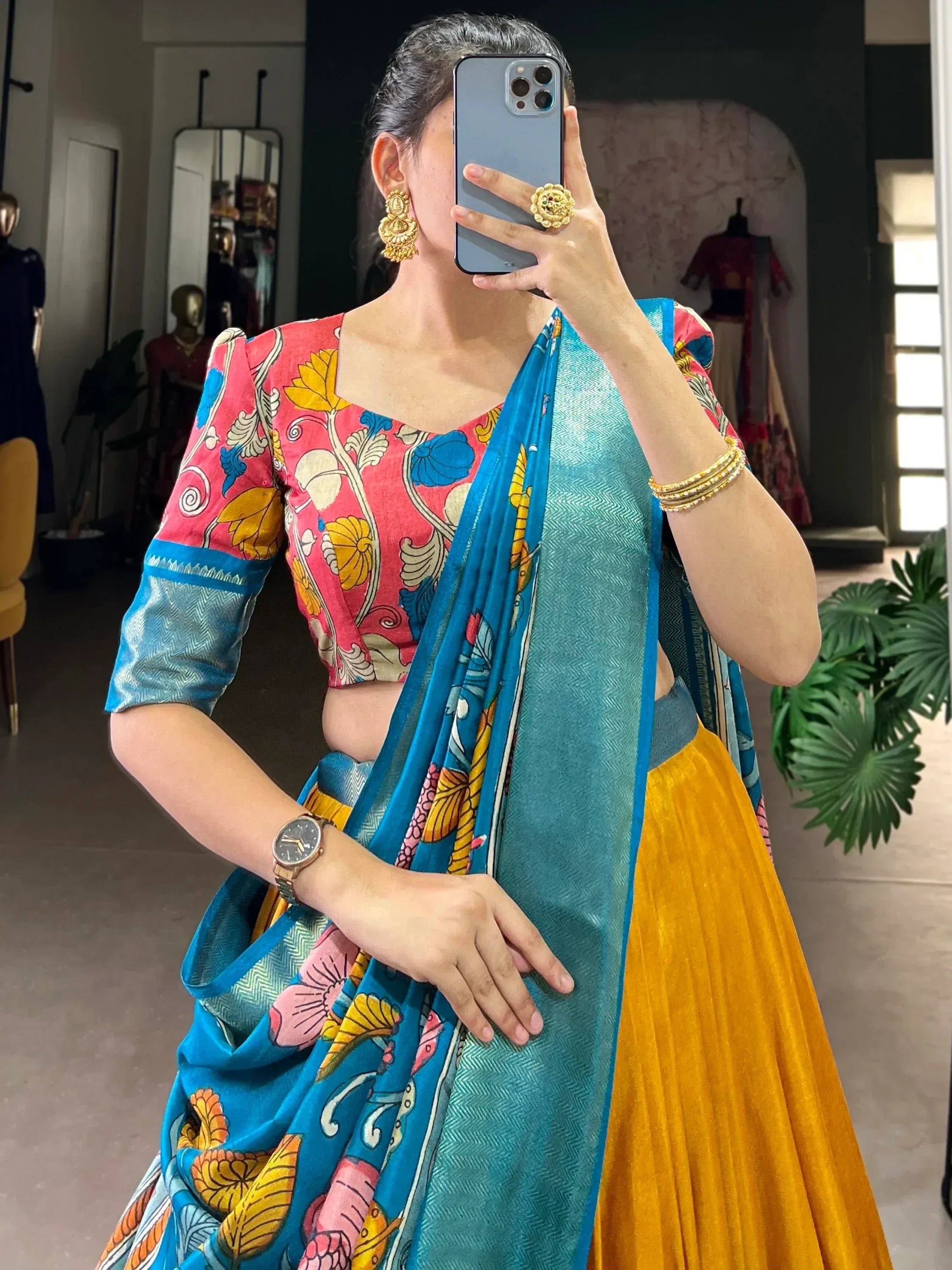 Woman in vibrant floral blouse and blue saree with yellow pleated skirt, styled with gold jewelry