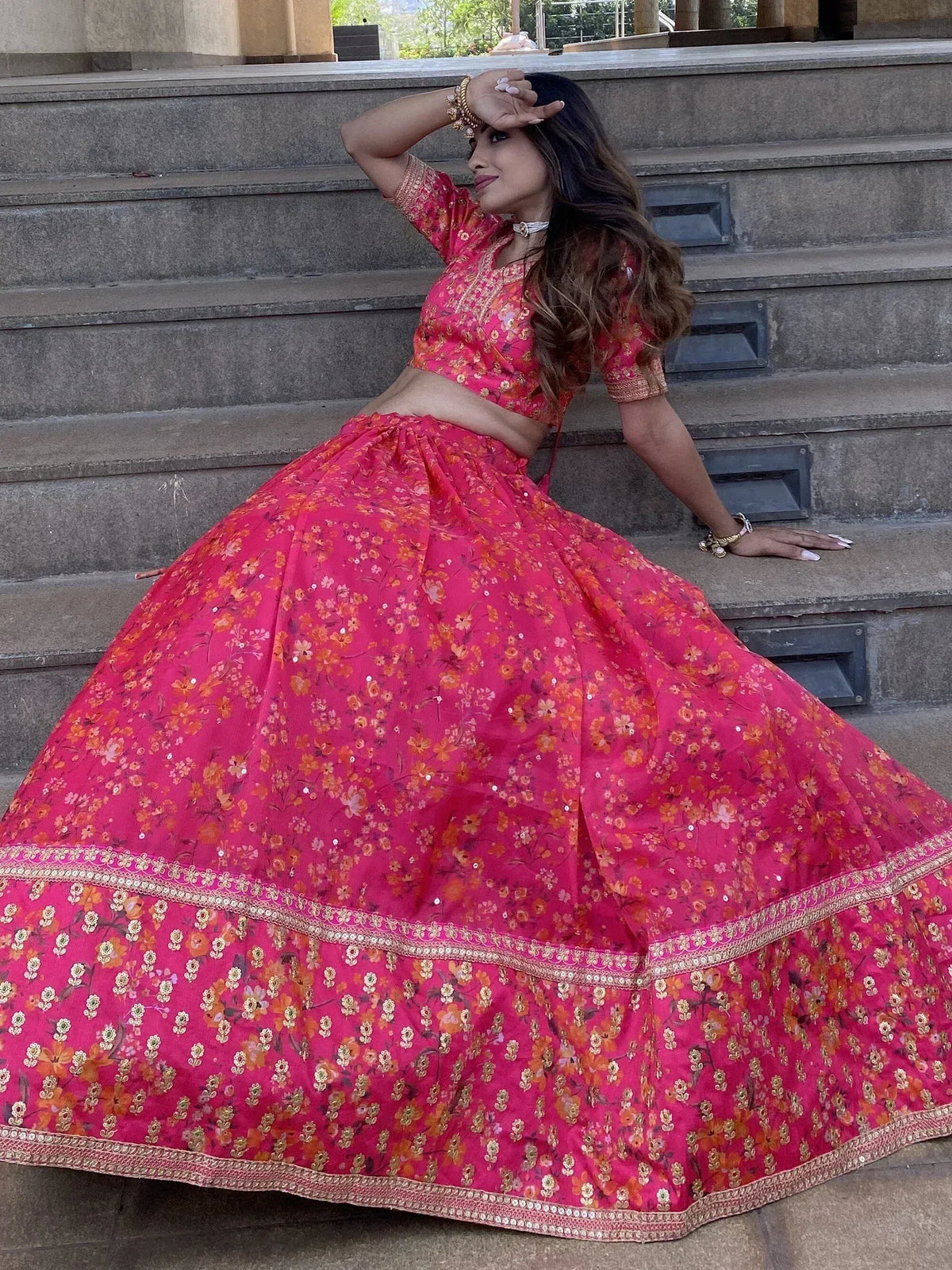 Woman in vibrant pink floral lehenga choli with gold embroidery, posing on stone steps