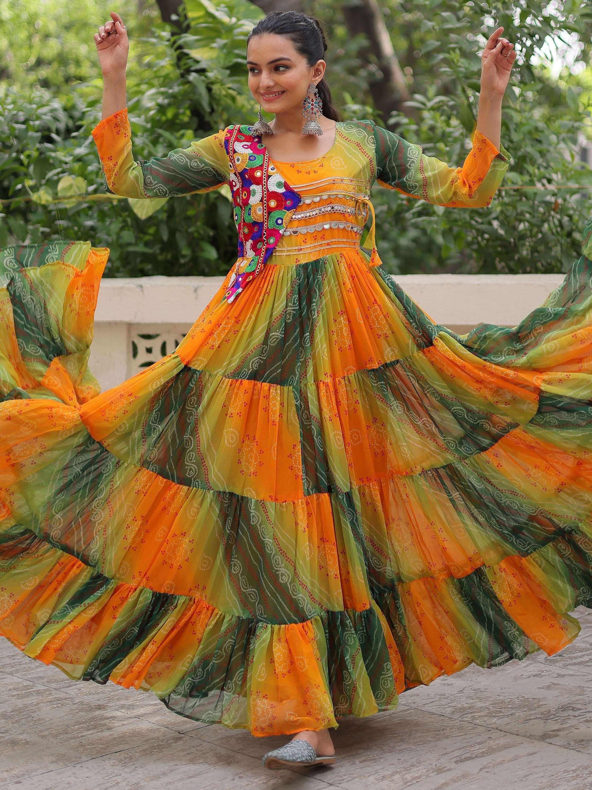 Woman in vibrant orange and green tiered ethnic gown with mirror work, dancing outdoors