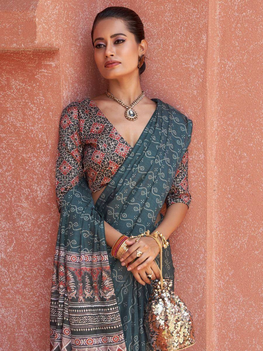 Woman in printed ethnic saree with floral blouse, gold jewelry, and sequin bag against textured wall