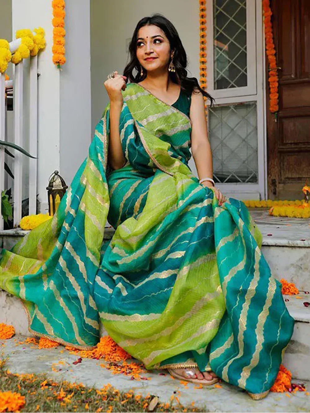 Woman in green and teal striped saree with gold trim, seated on steps adorned with marigold flowers