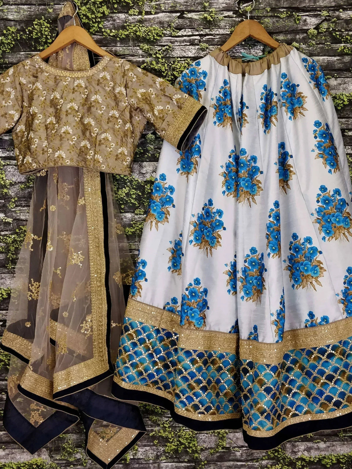 Blue floral and gold embroidered lehenga choli sets with sheer dupatta, hanging on wooden hangers against rustic wall