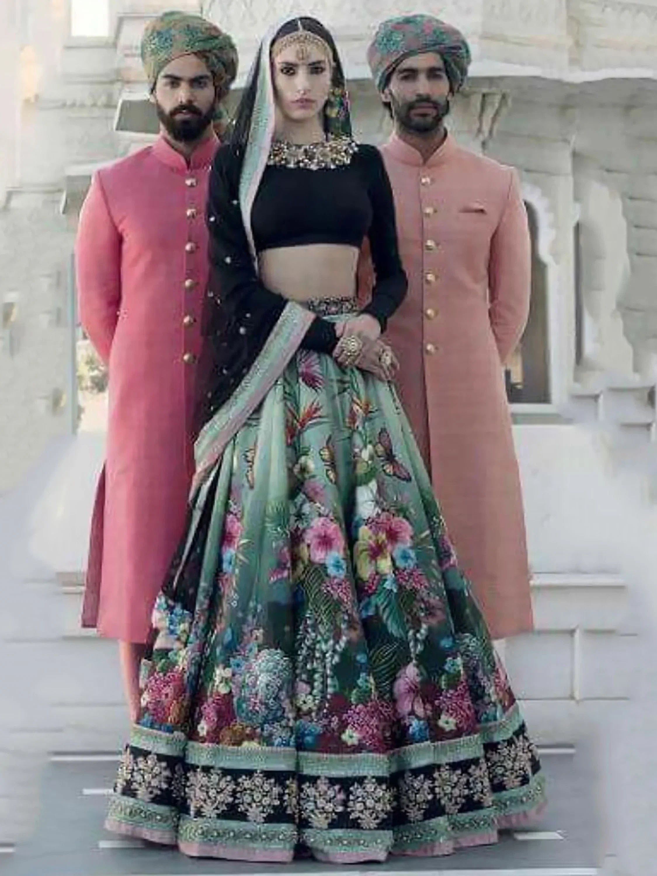 Woman in floral lehenga choli with black blouse, flanked by men in pink sherwanis and turbans, Indian ethnic wear