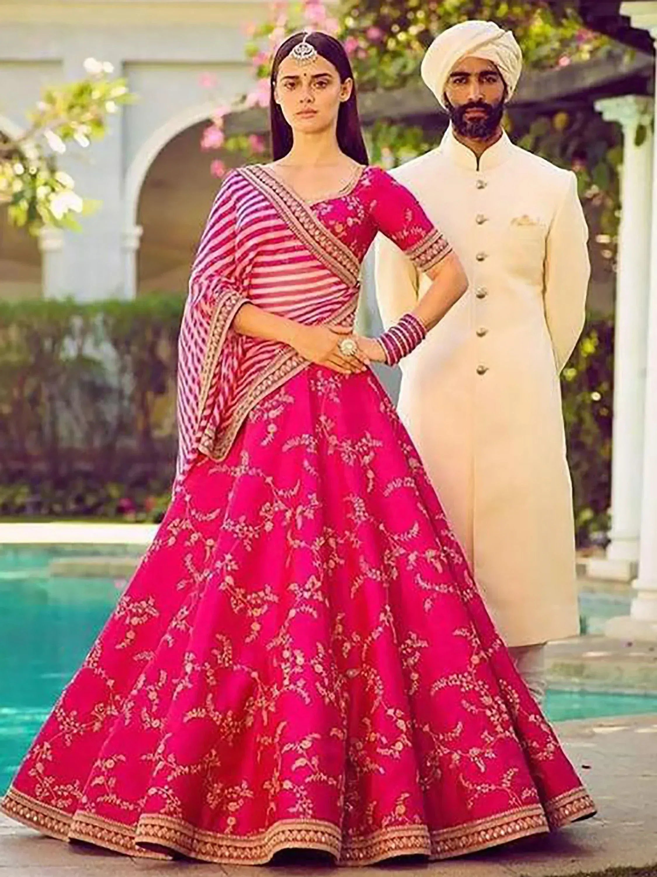Woman in pink embroidered lehenga choli with man in cream sherwani, garden setting