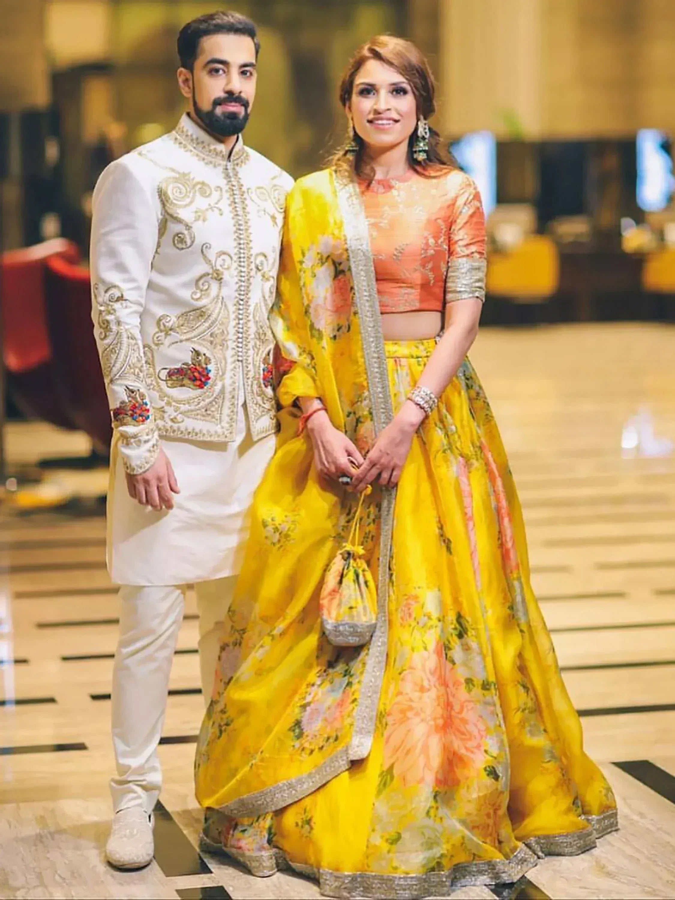 Couple in Indian wedding attire: woman in yellow floral lehenga choli, man in white embroidered sherwani