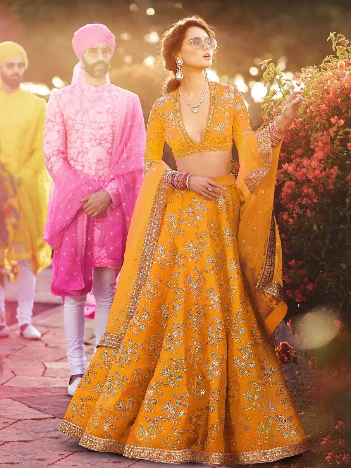 Woman in vibrant yellow embroidered lehenga choli with men in pink and yellow sherwani, garden setting