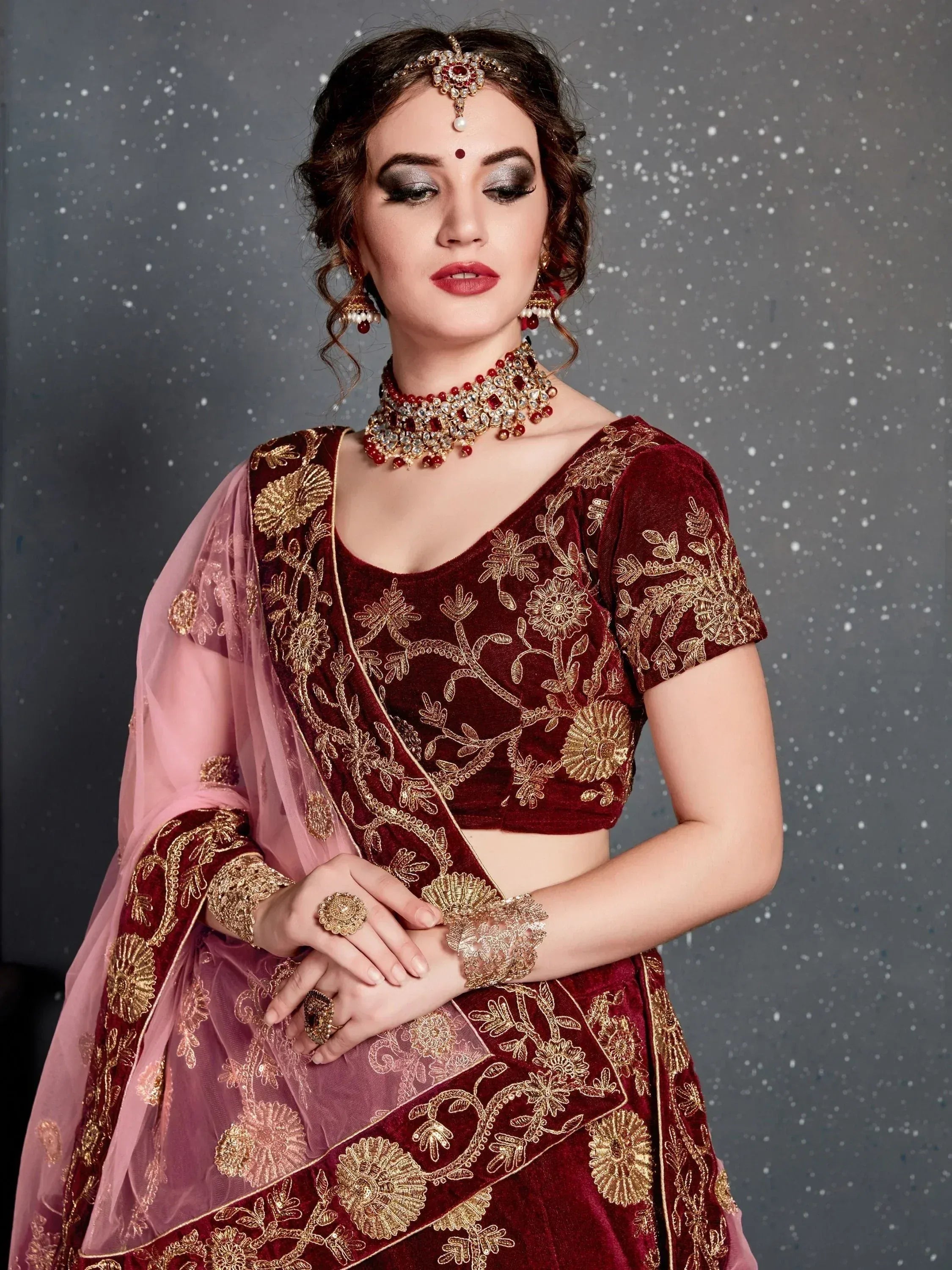 Maroon velvet lehenga choli with gold embroidery, pink dupatta, and traditional Indian jewelry for weddings