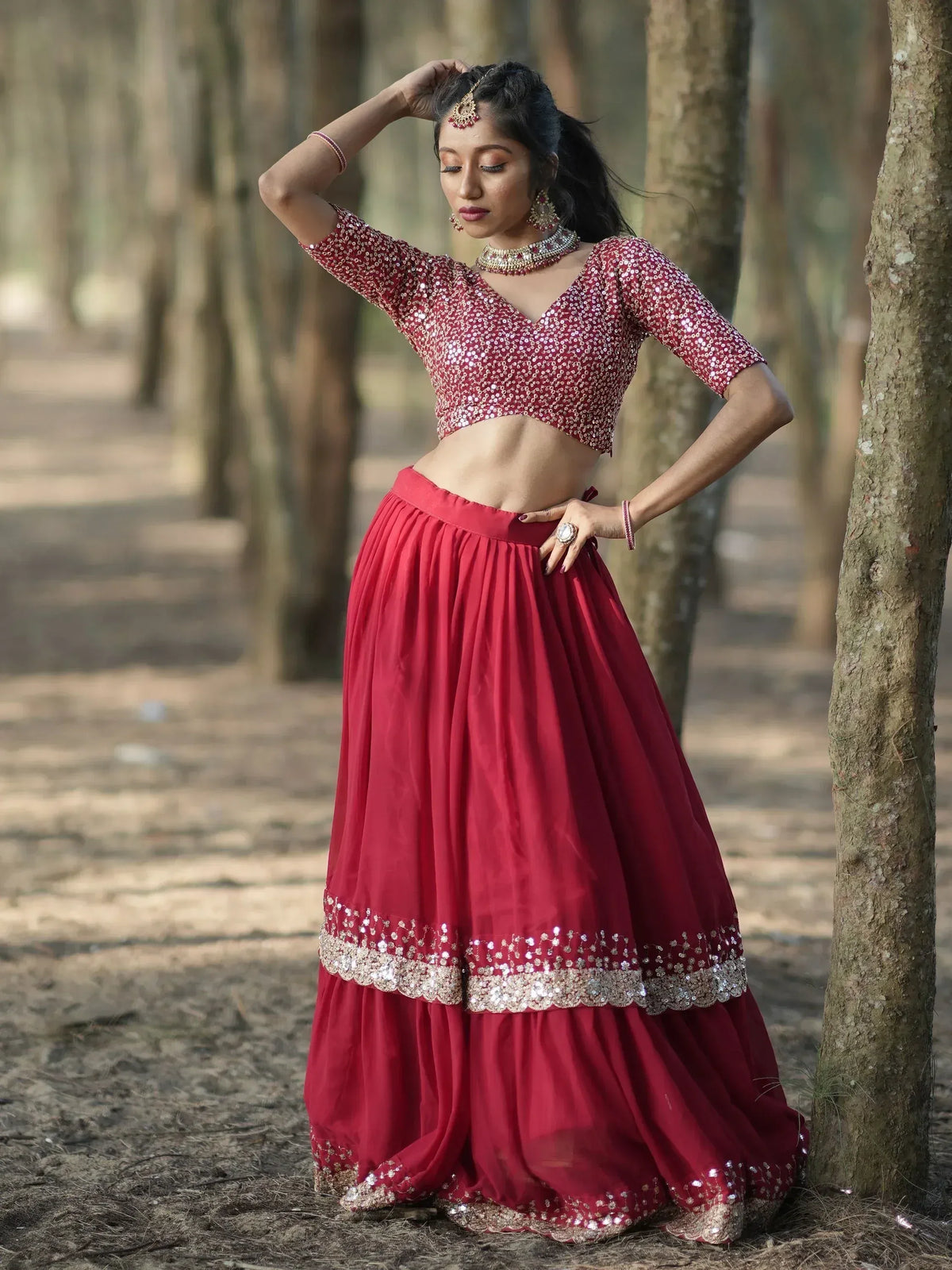 Woman in maroon sequin lehenga choli with gold jewelry, posing in forest setting