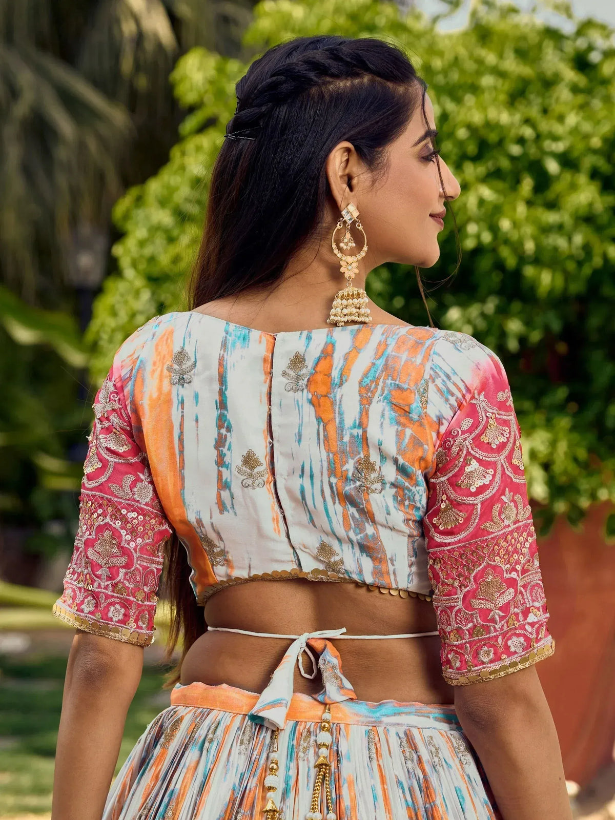 Woman in colorful printed lehenga choli with embroidered sleeves and gold jhumka earrings, outdoor garden setting