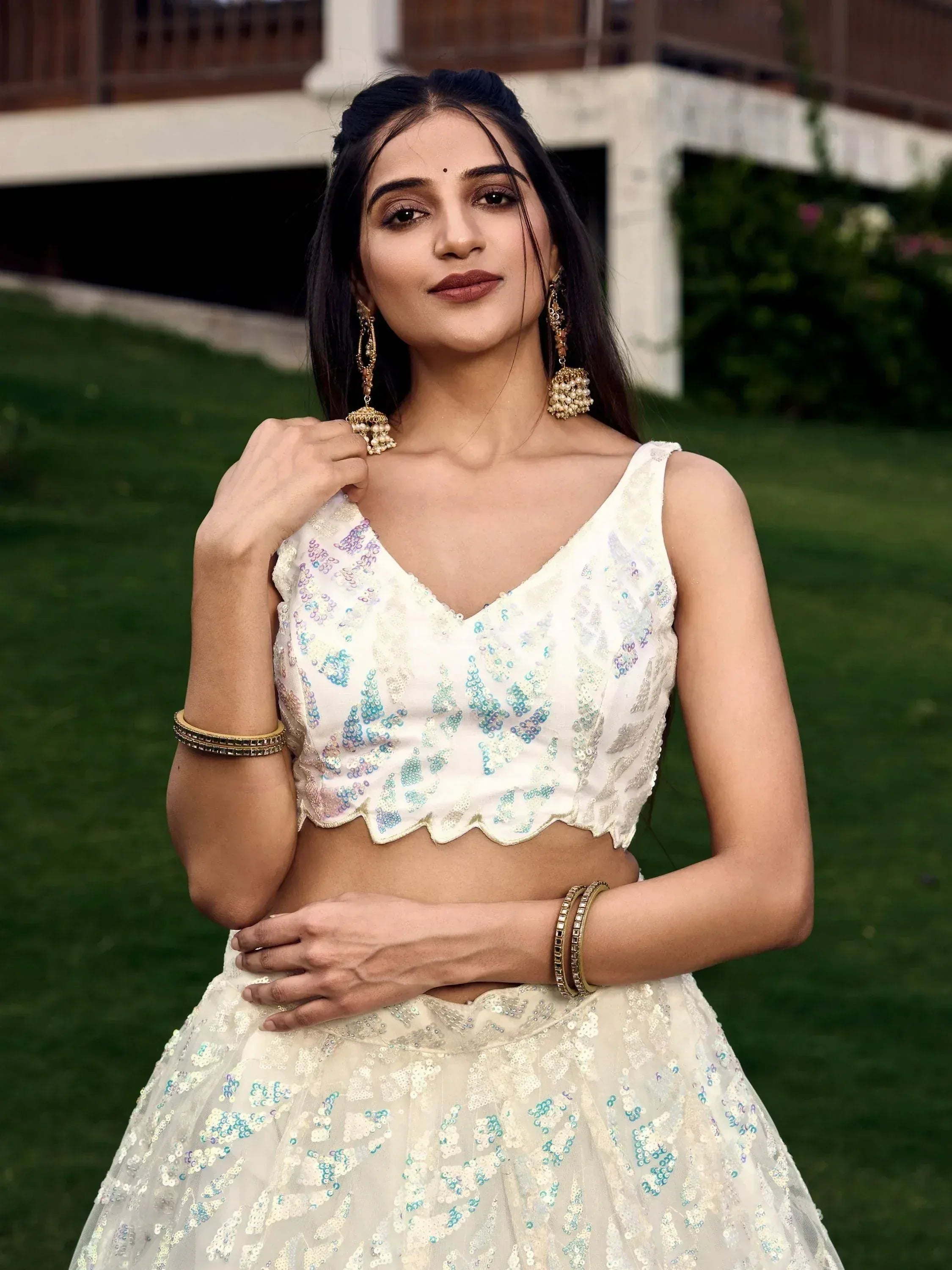 Woman in white sequined lehenga choli with blue floral embroidery, gold jhumka earrings, outdoor garden setting