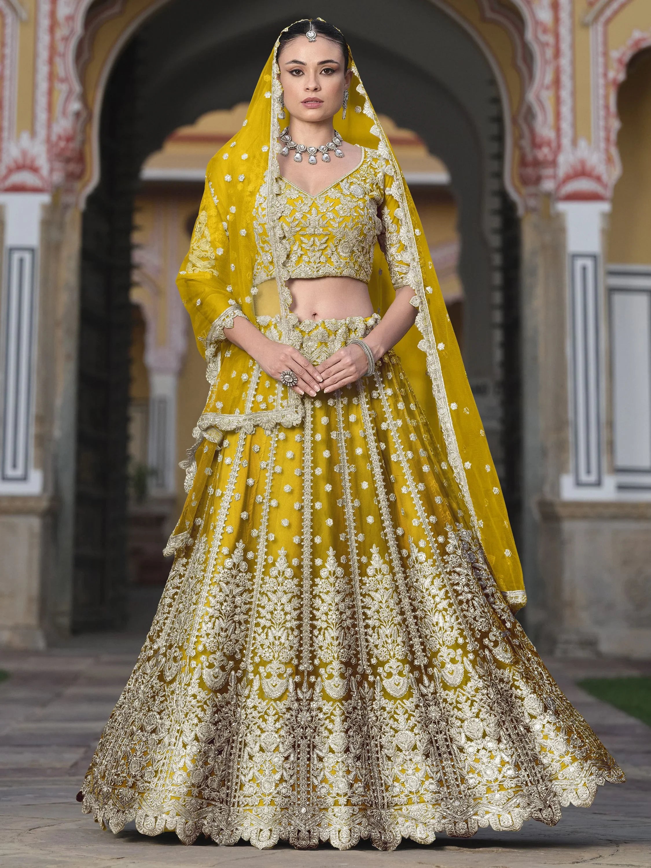 Woman in yellow bridal lehenga choli with intricate embroidery, standing in traditional courtyard