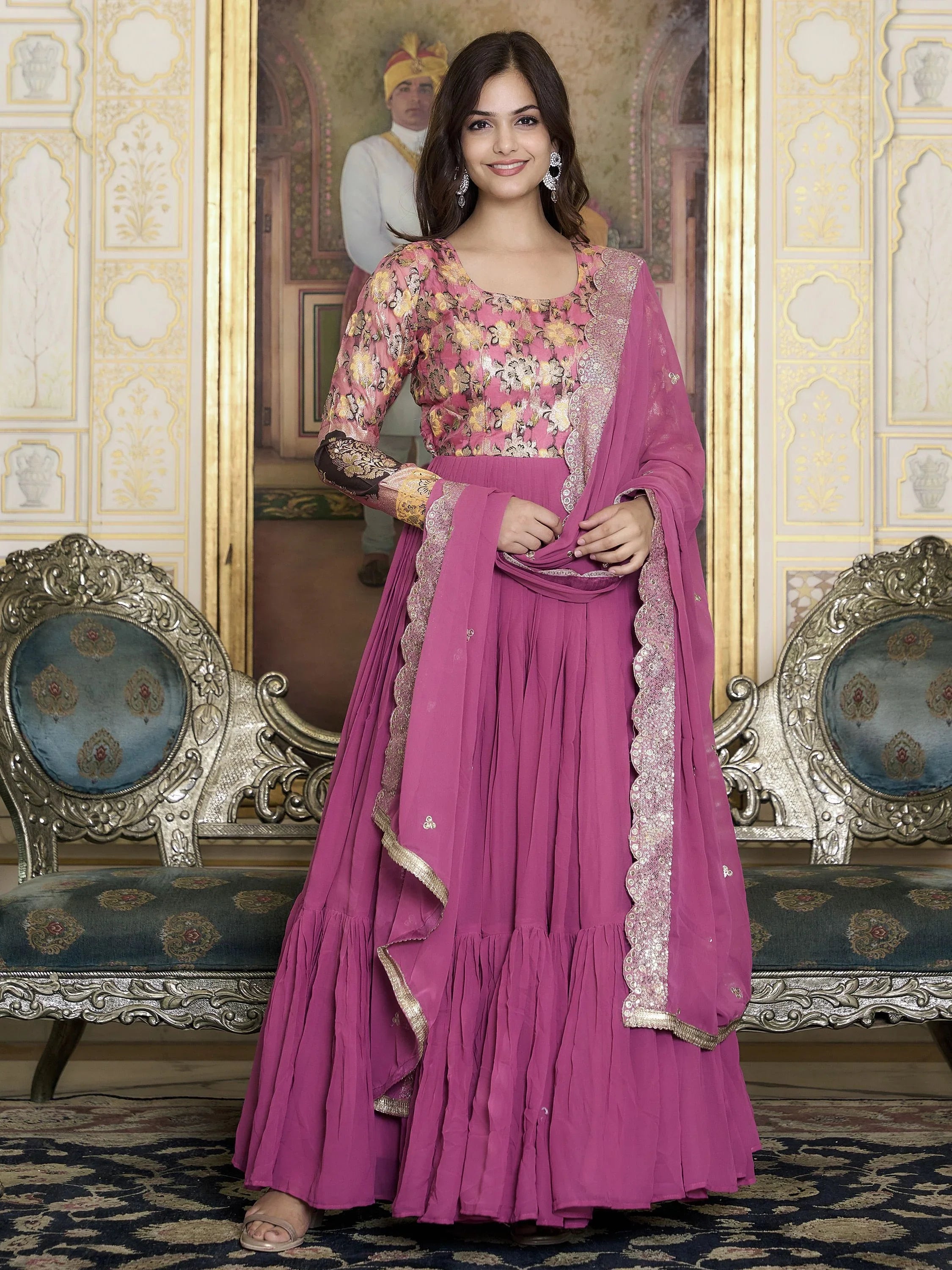 Woman wearing a pink embroidered ethnic gown with dupatta, standing in a royal-themed room.