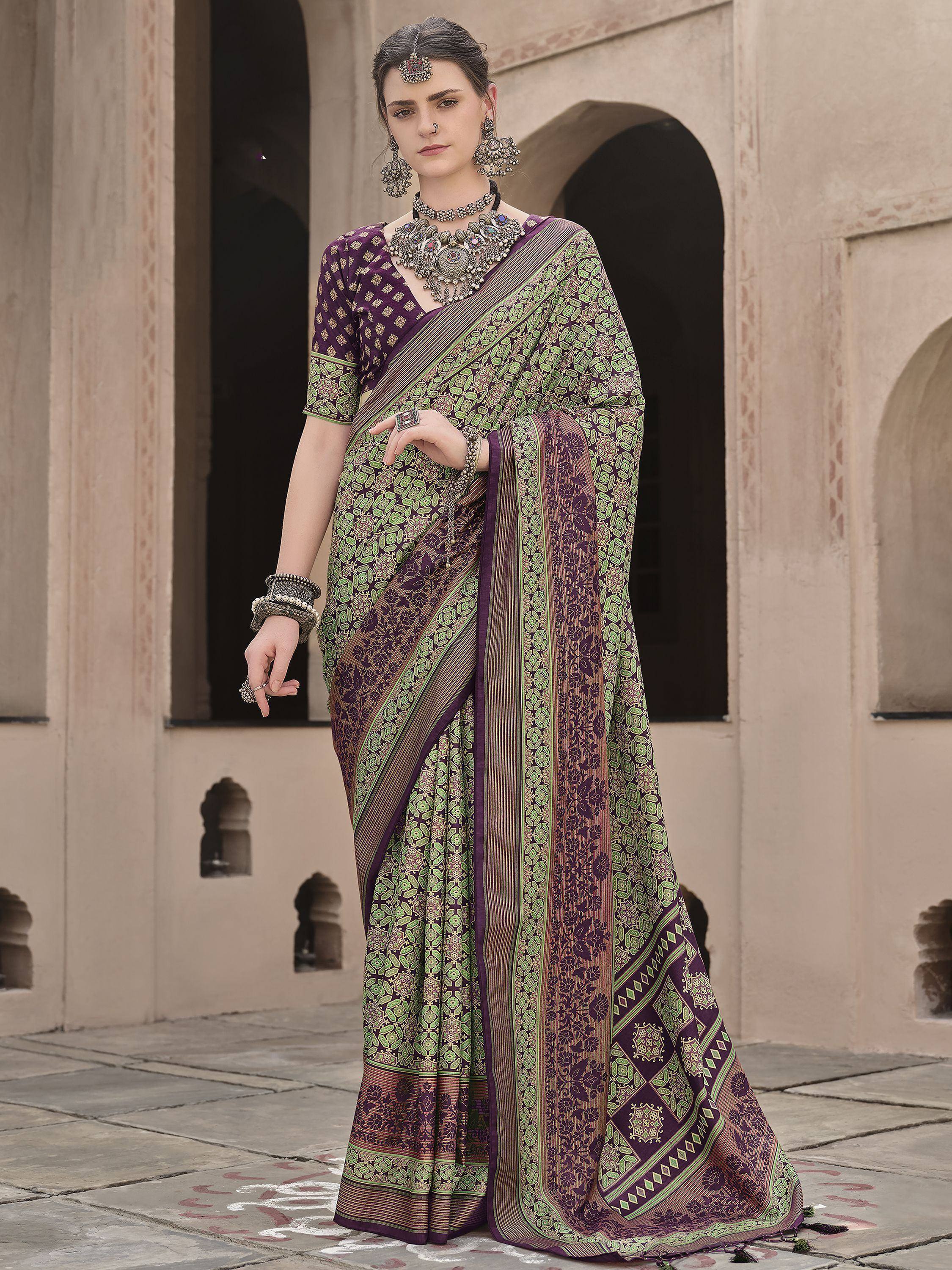 Woman in purple and green printed saree with traditional jewelry, standing in courtyard