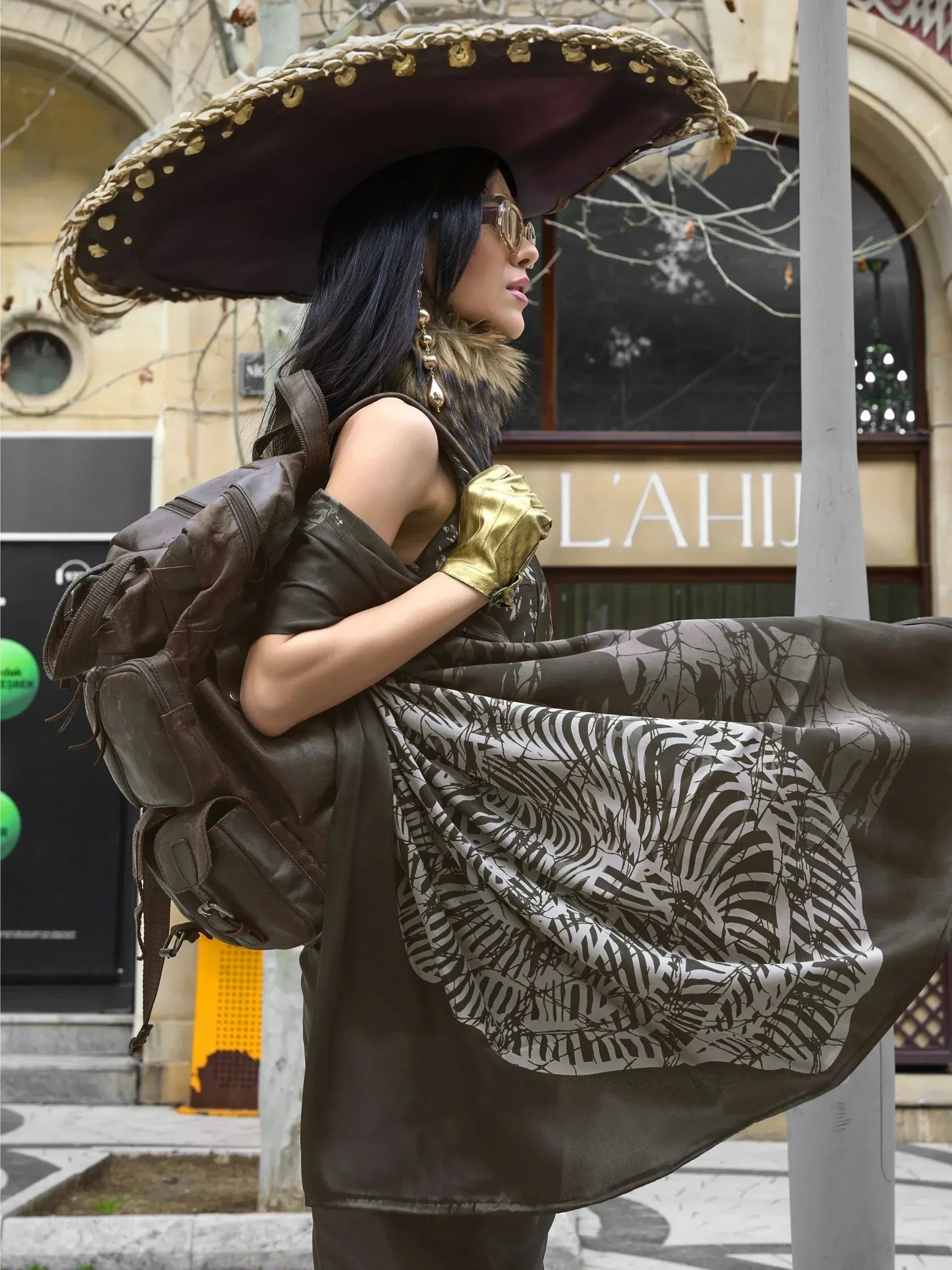 Woman in ethnic print gown with wide-brimmed hat, gold gloves, and backpack, street style fashion