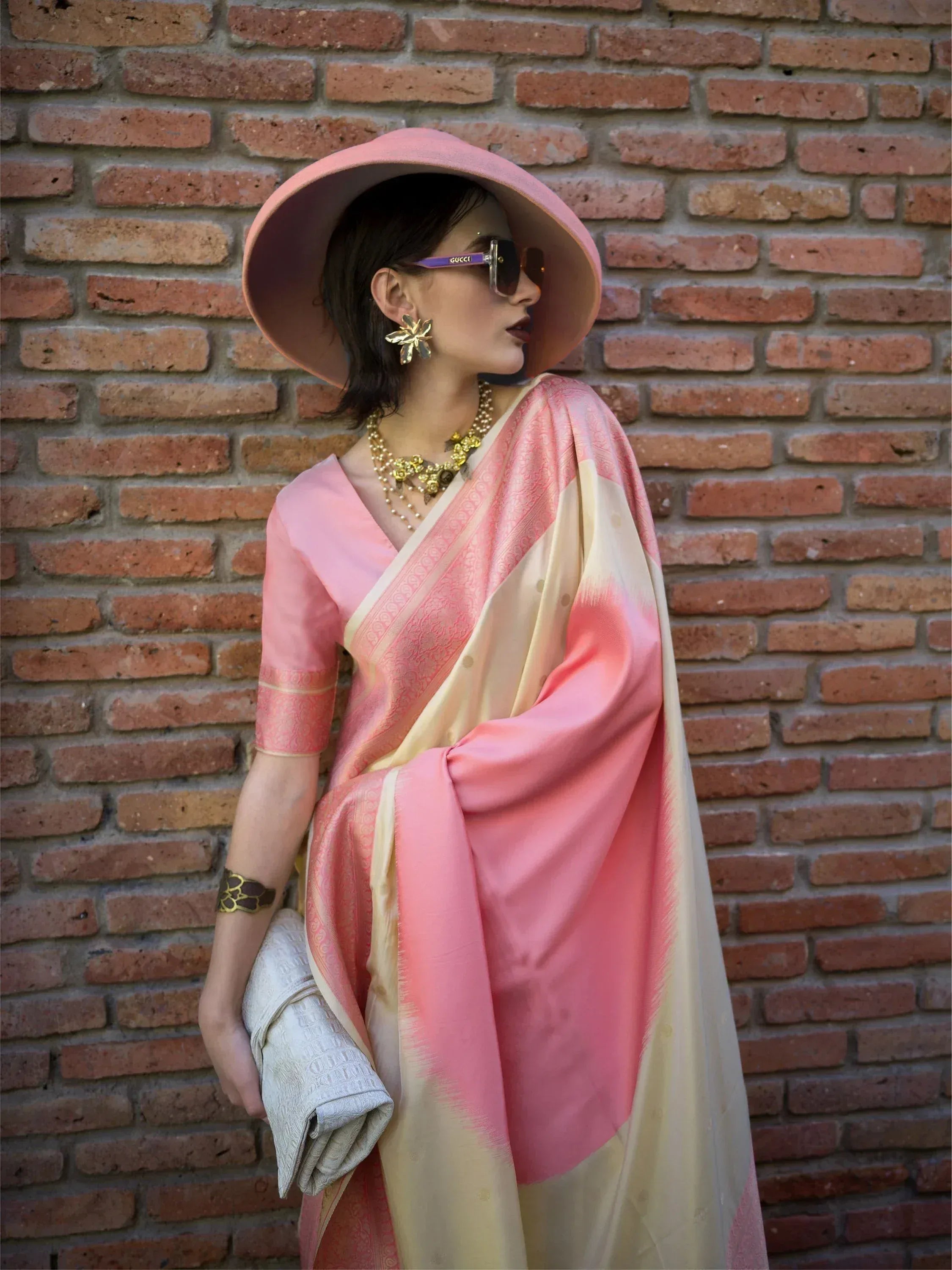 Woman in pink and cream designer saree with gold jewelry, wide-brim hat, against brick wall