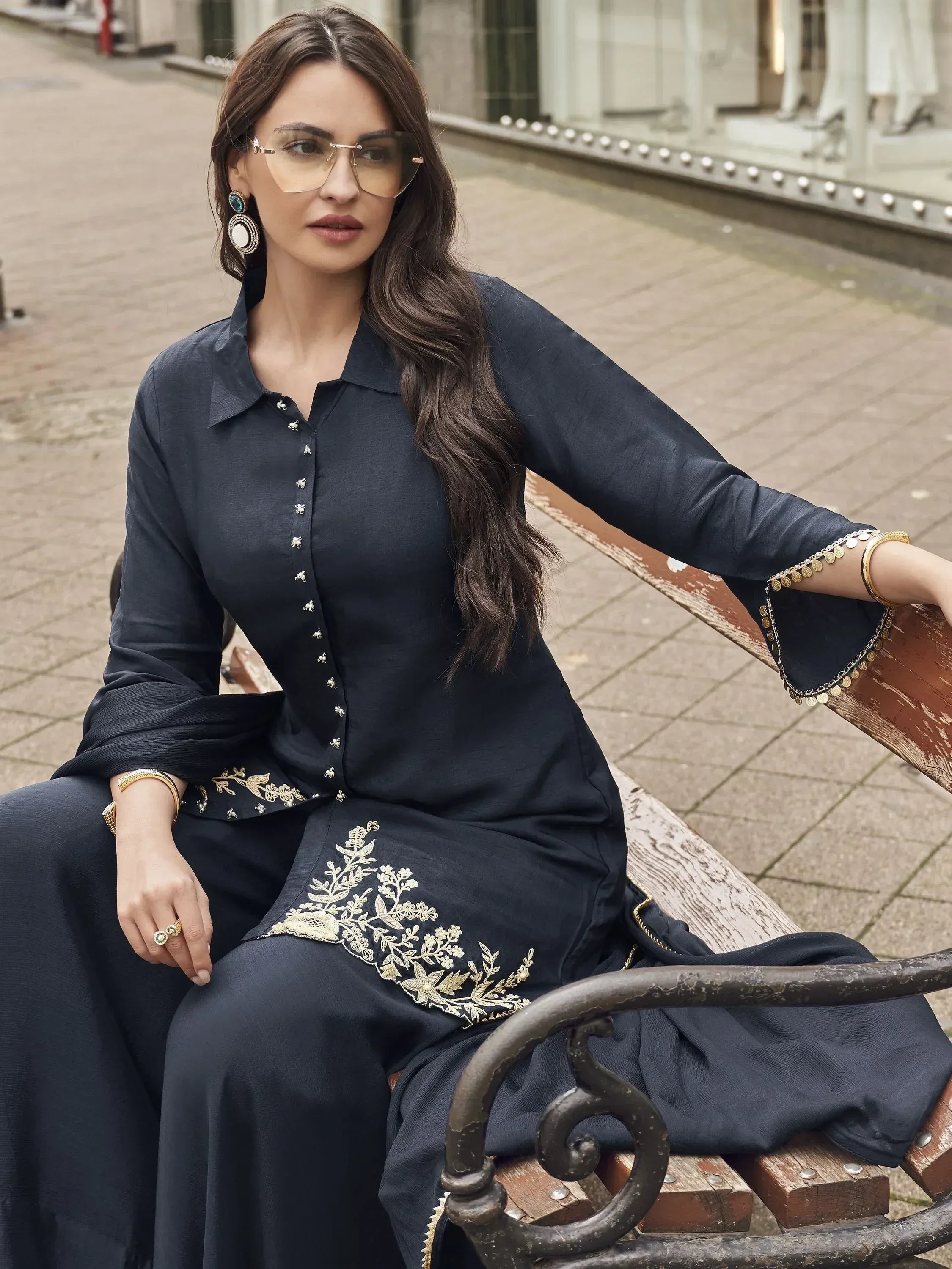 Woman in navy blue embroidered salwar kameez with gold detailing, sitting on bench outdoors