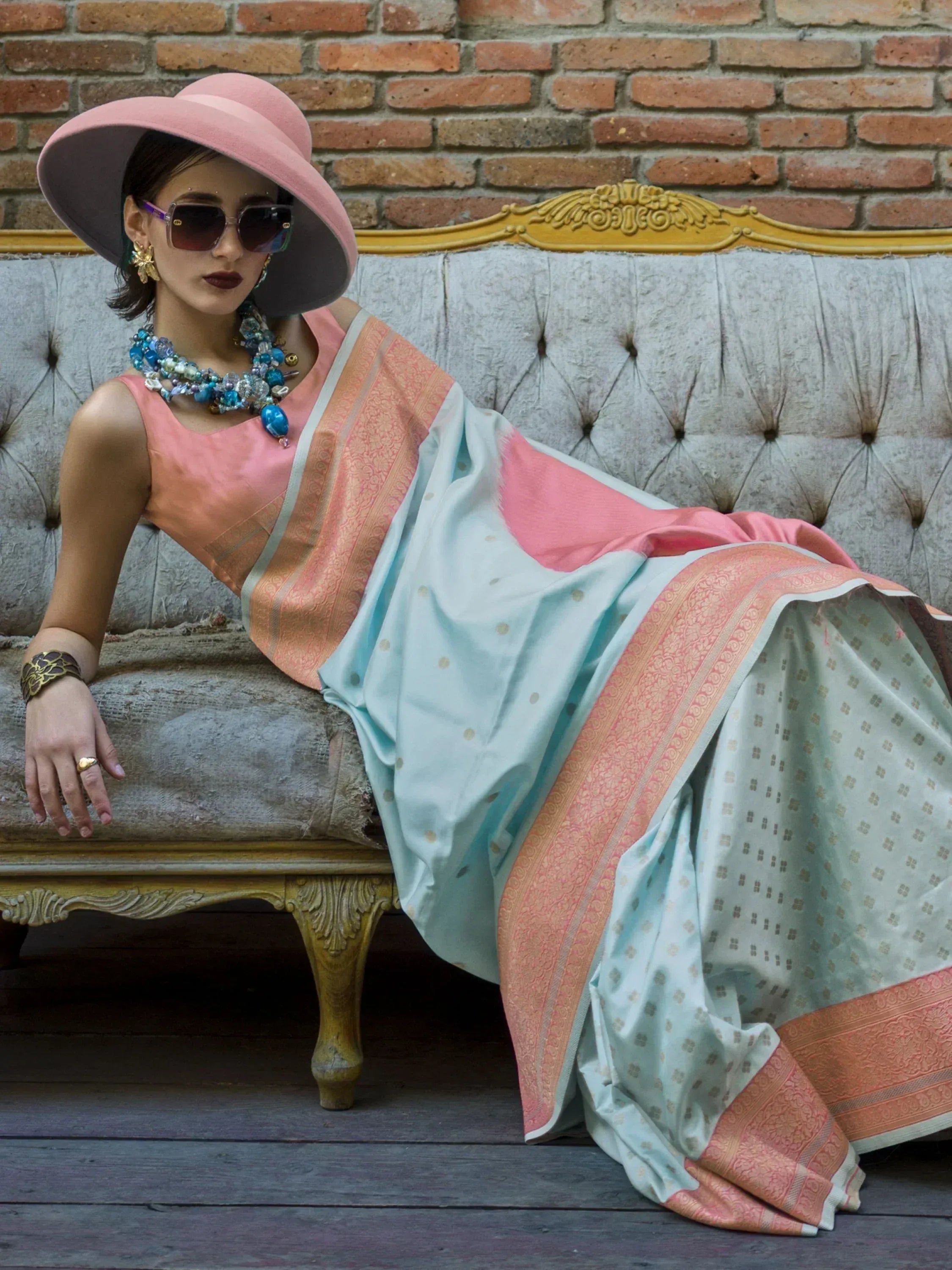 Woman in pastel blue and pink designer saree with gold border, styled with wide-brim hat and statement jewelry