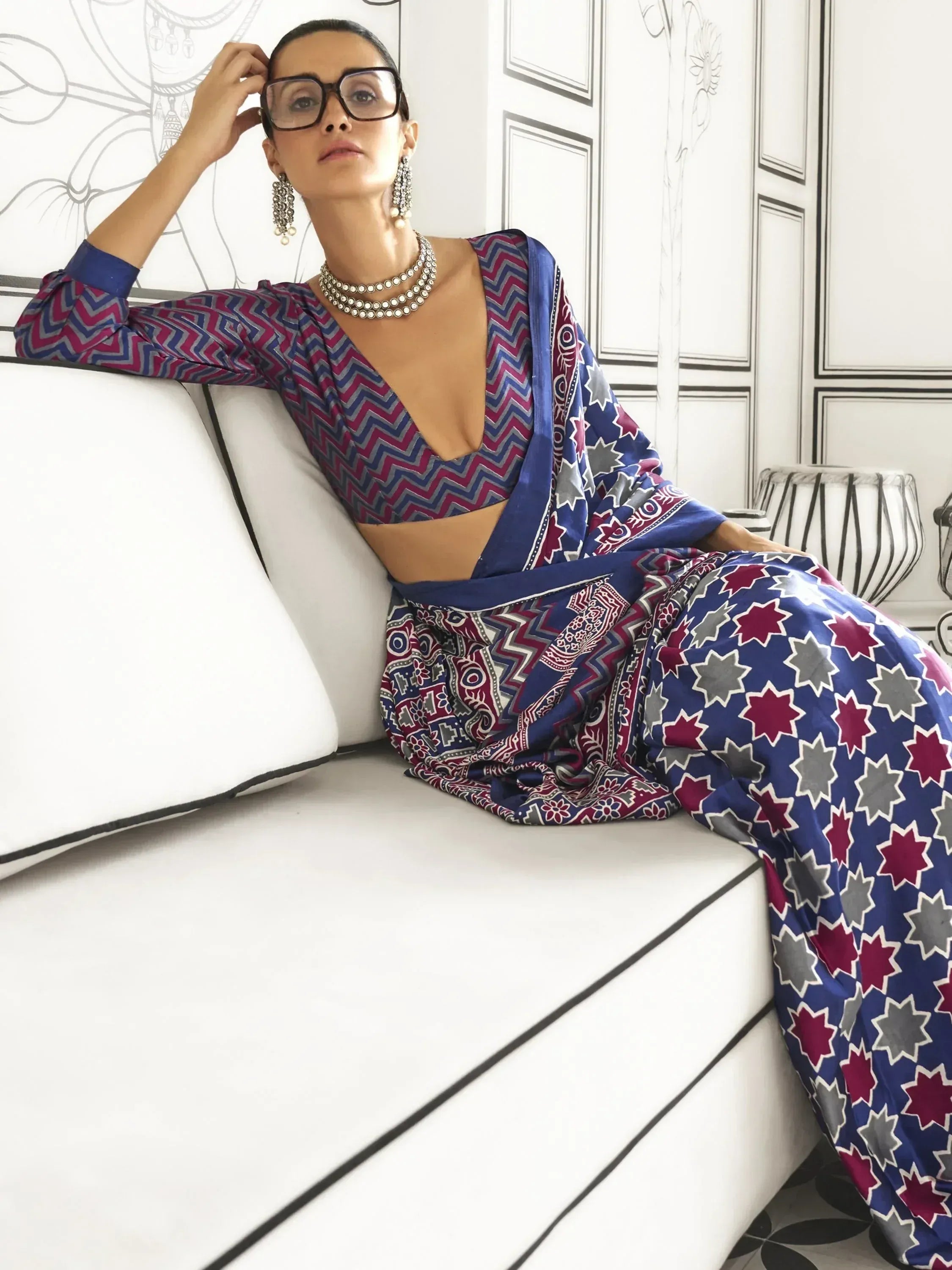 Woman in vibrant ethnic saree with geometric print, styled with statement jewelry and glasses, seated on white sofa.