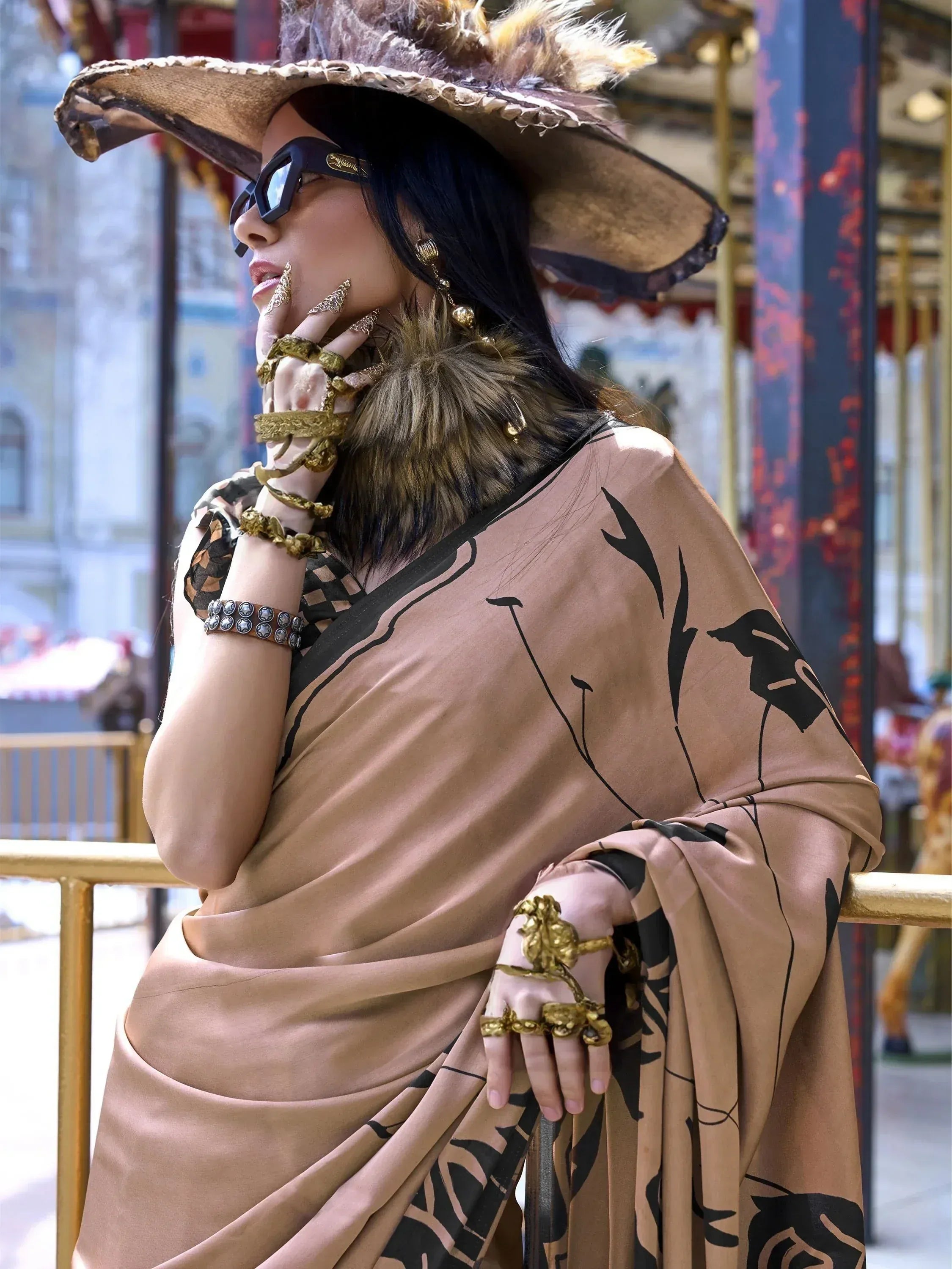 Woman in beige saree with black floral print, wide-brimmed hat, and gold jewelry, styled for ethnic fashion