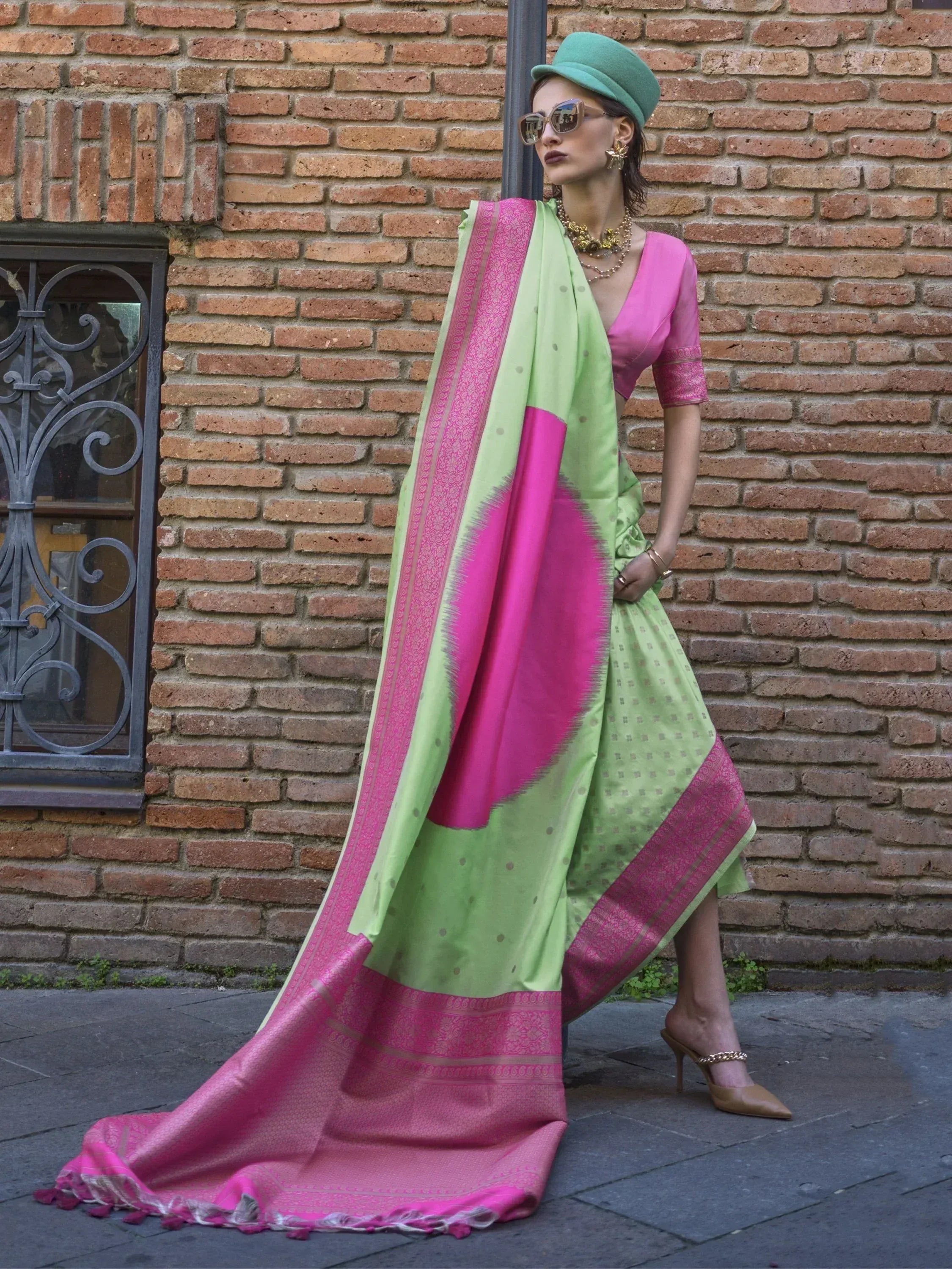 Woman in vibrant green and pink saree with designer blouse, styled with hat and sunglasses against brick wall