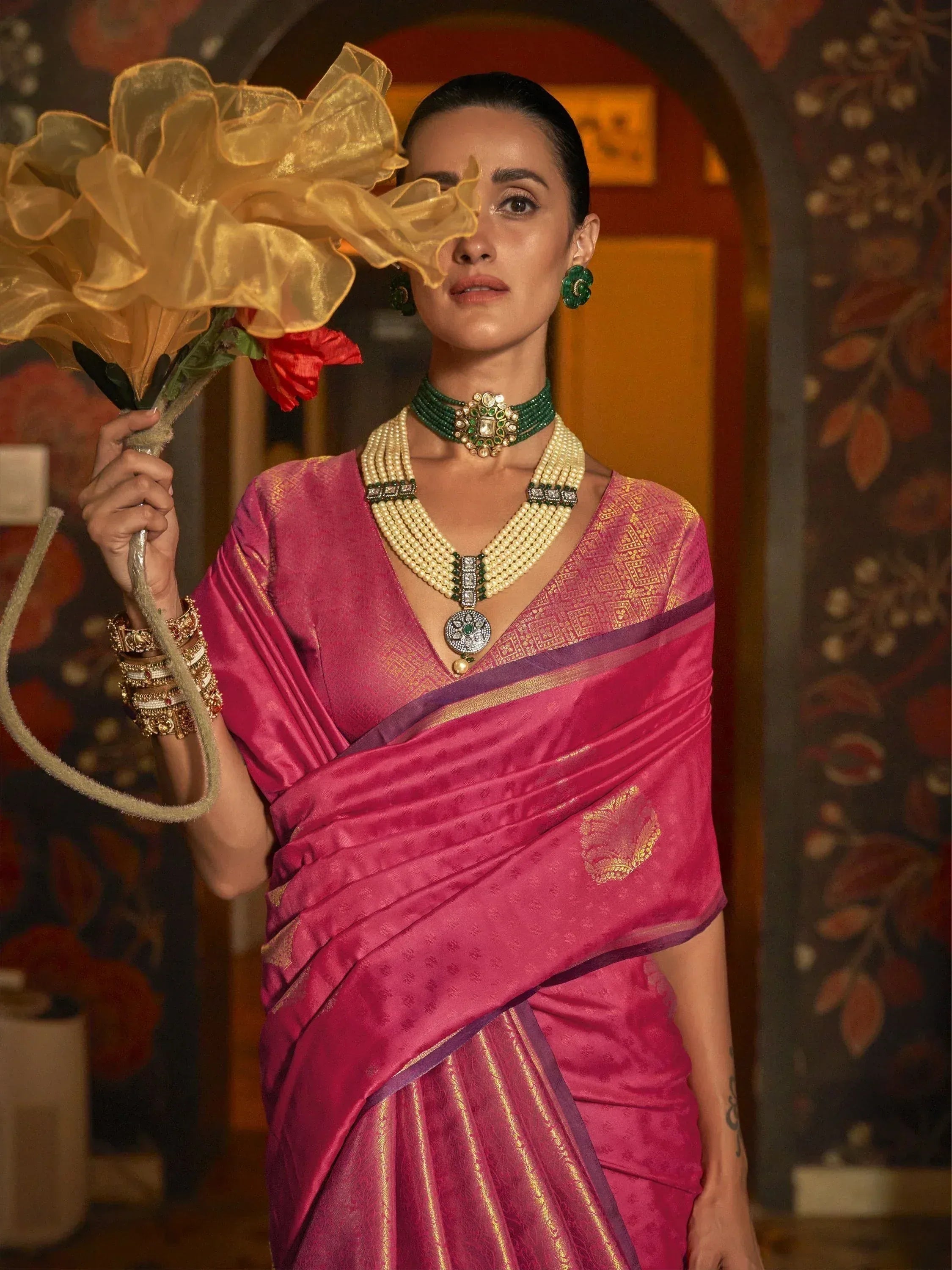 Woman in pink silk saree with gold embroidery, green jewelry, holding golden flower, ethnic Indian wear
