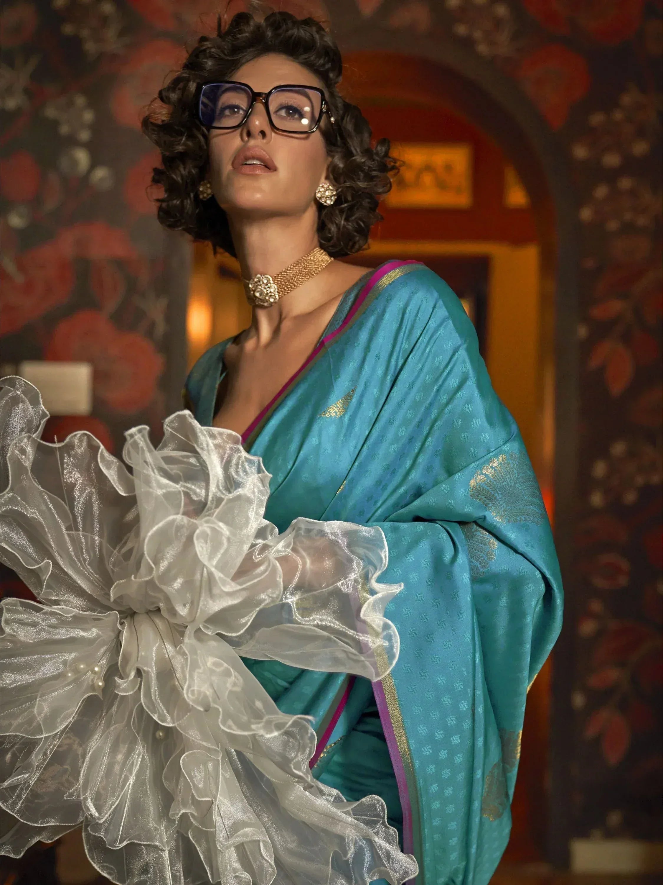 Stylish woman in teal designer saree with ruffled blouse, gold jewelry, and glasses