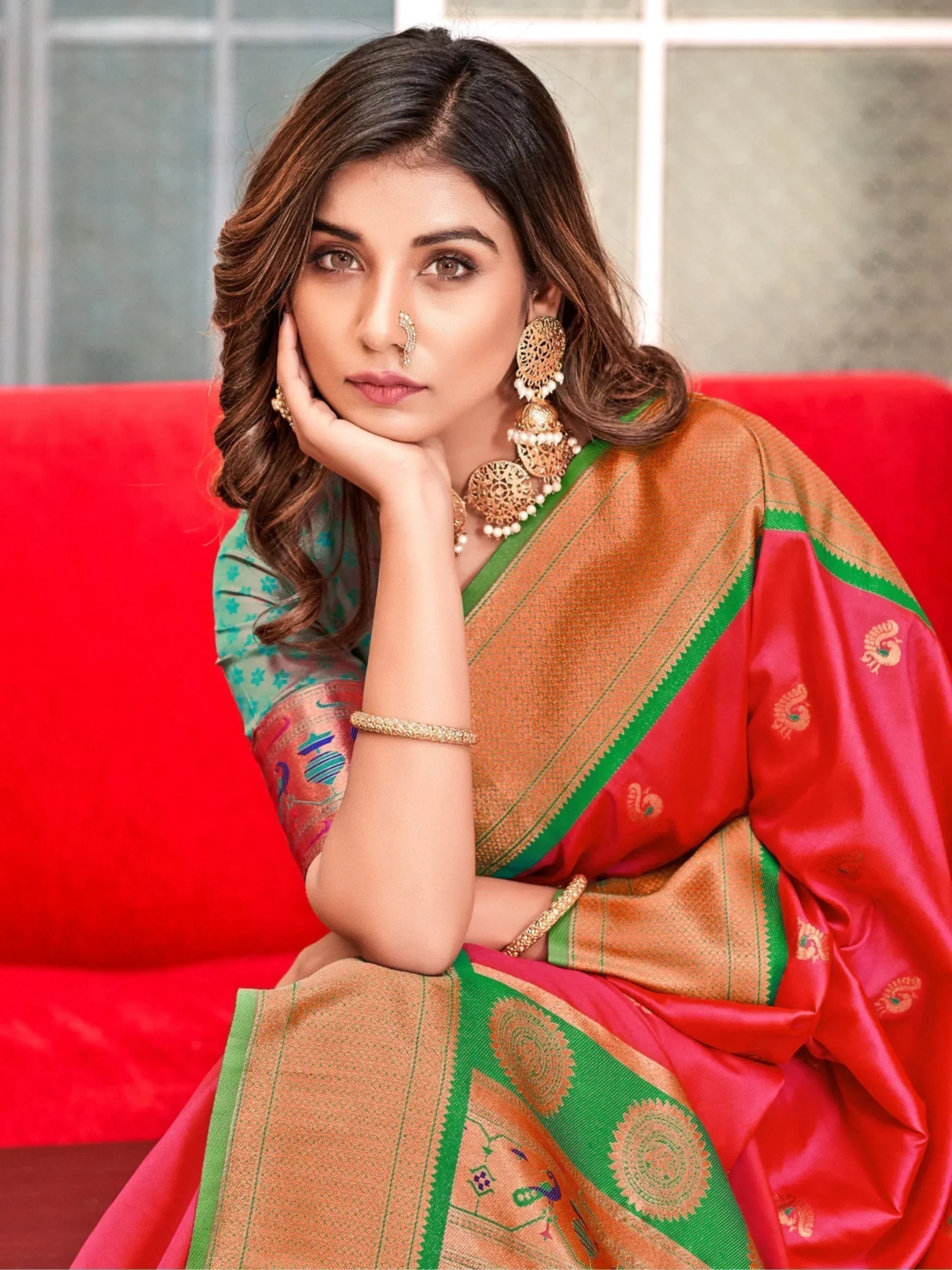 Woman in vibrant red and gold Indian saree with green border, traditional jewelry, sitting on red sofa
