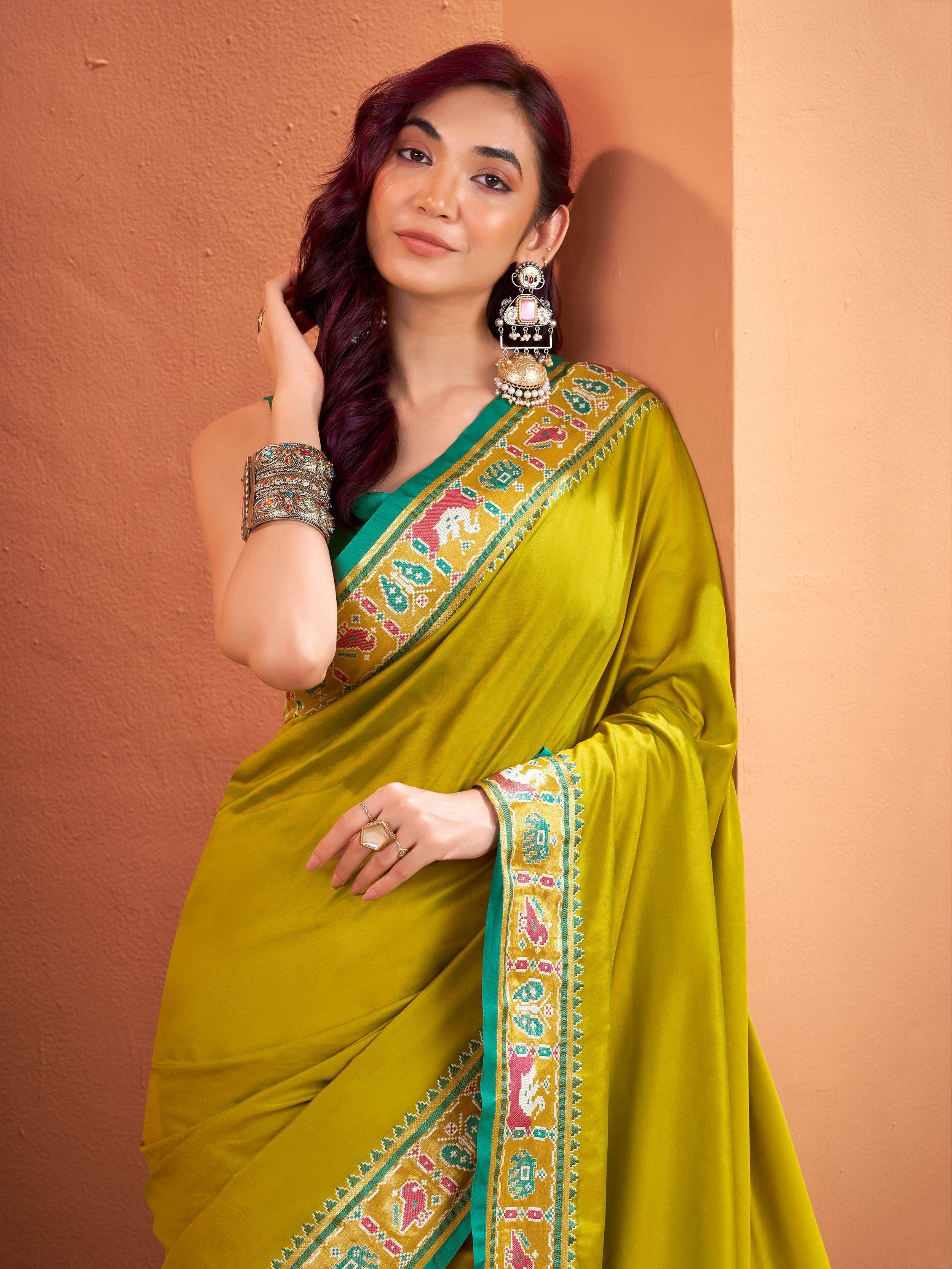 Woman in vibrant green saree with embroidered border, ethnic jewelry, and purple hair against warm wall