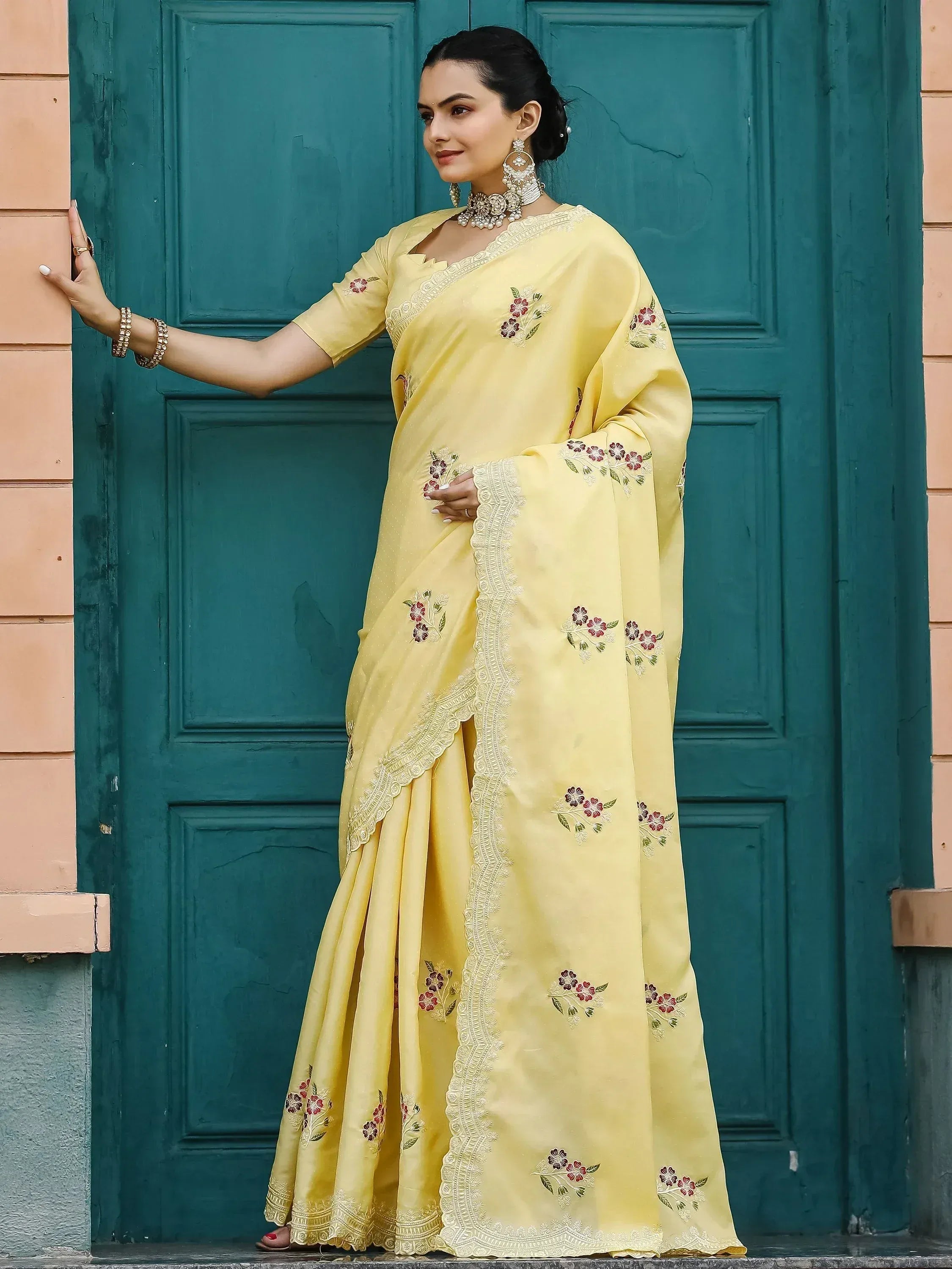 Yellow floral embroidered saree with lace border, styled with traditional jewelry against teal door