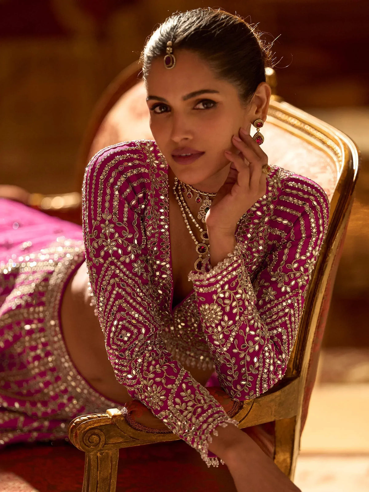 Woman in pink designer lehenga choli with mirror work, seated on ornate chair, wearing traditional Indian jewelry