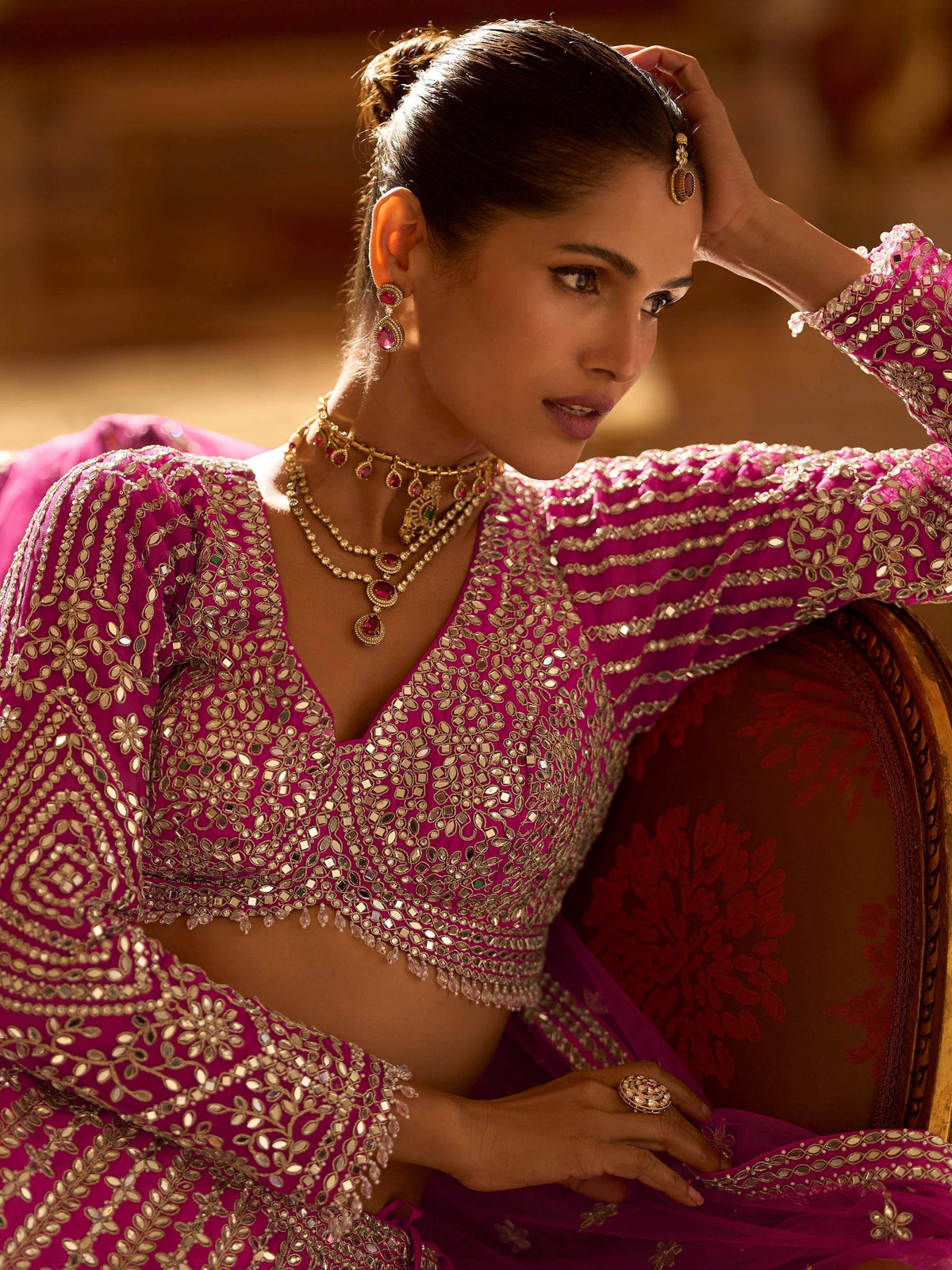 Elegant woman in magenta mirror-work lehenga choli with gold jewelry, showcasing Indian ethnic wear.