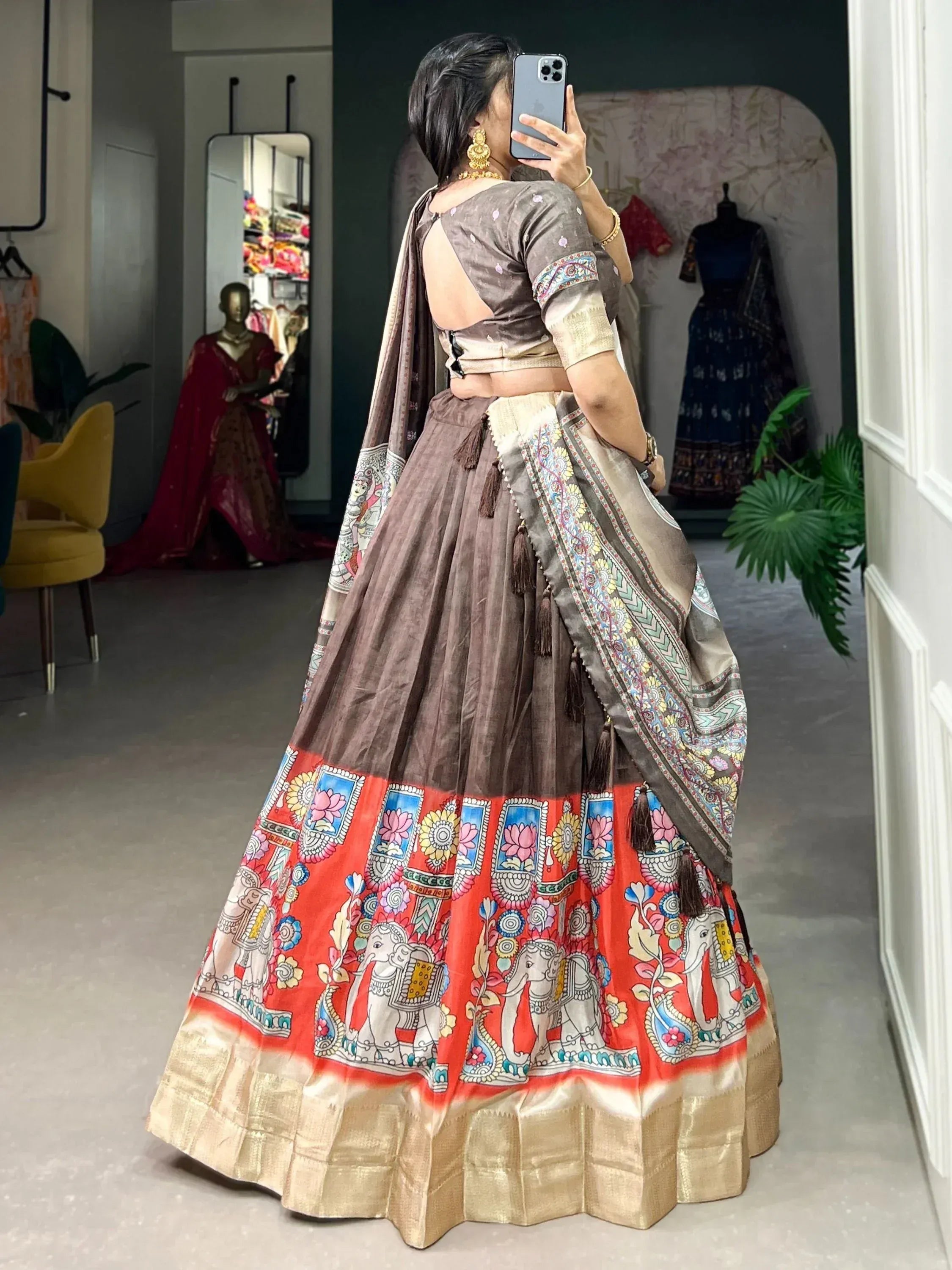 Woman wearing brown and red printed lehenga choli with elephant motifs, taking mirror selfie in boutique