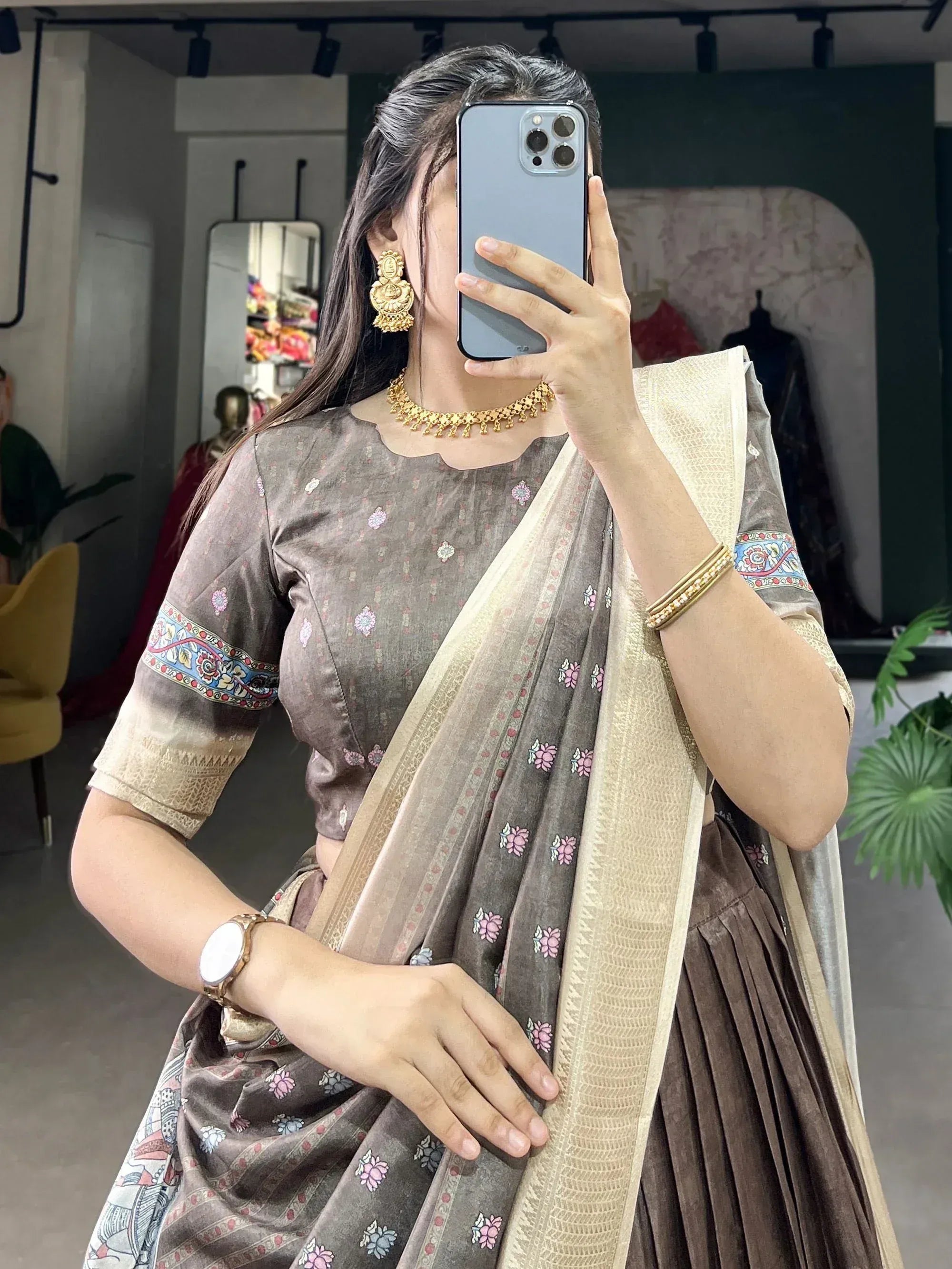 Woman in brown floral printed saree with gold jewelry, taking mirror selfie in ethnic wear store