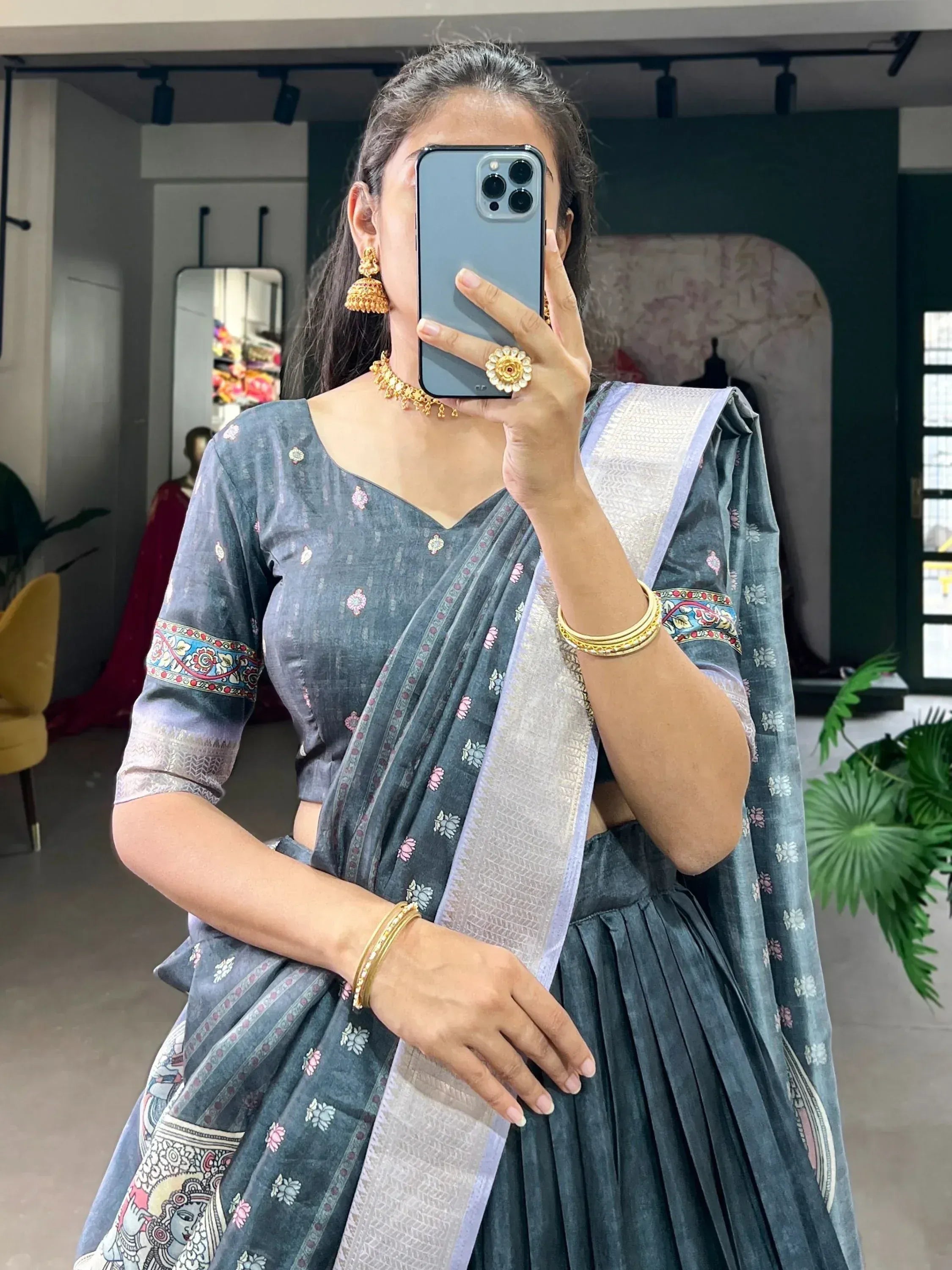 Woman in grey floral saree with silver border, gold jewelry, taking mirror selfie in boutique