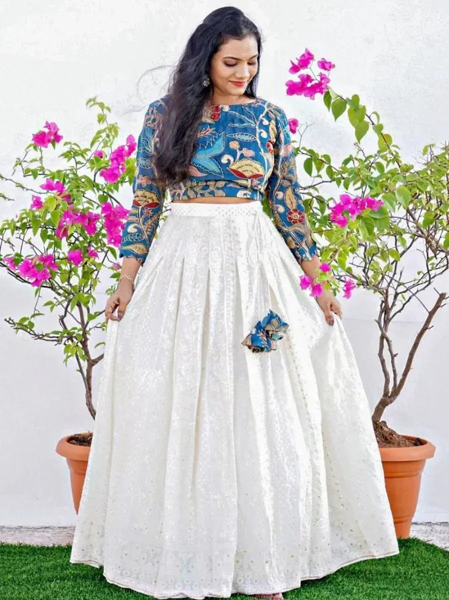 Woman in blue floral crop top and white embroidered lehenga skirt, standing by pink flowers