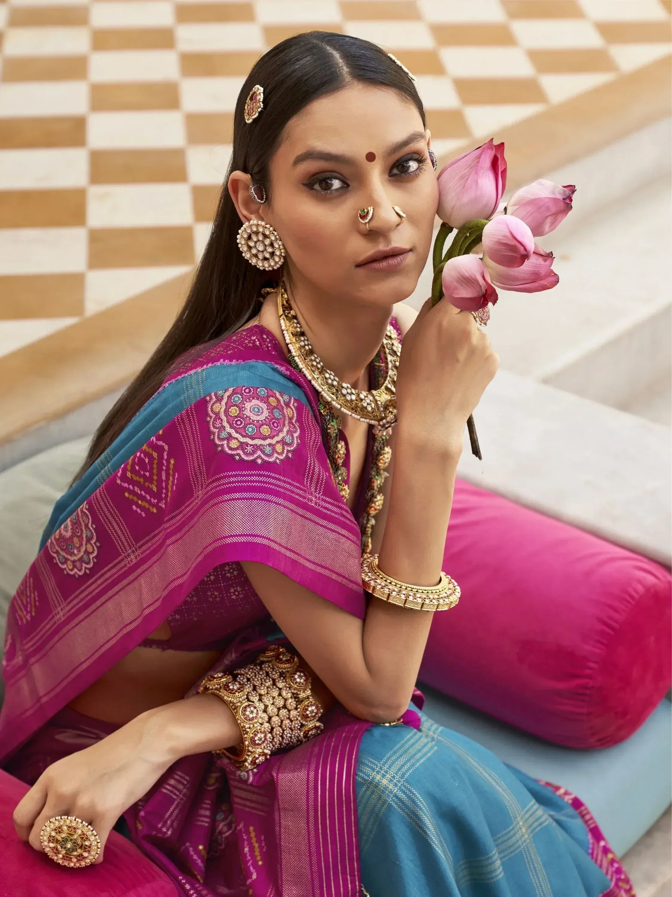 Woman in vibrant purple and blue ethnic saree with intricate embroidery, traditional jewelry, holding pink lotus flowers