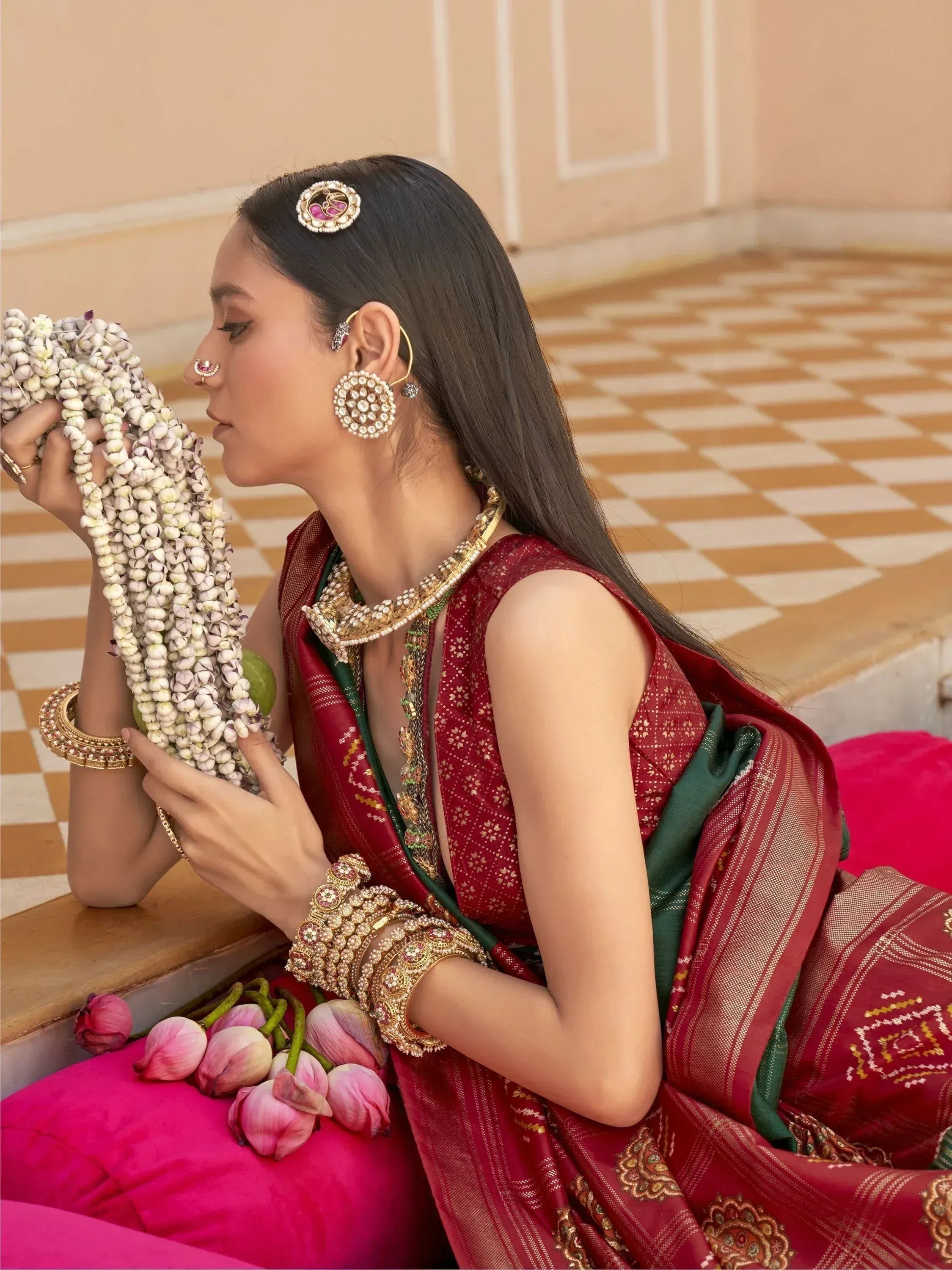 Woman in red and green ethnic saree with gold jewelry, holding floral garland, traditional Indian attire