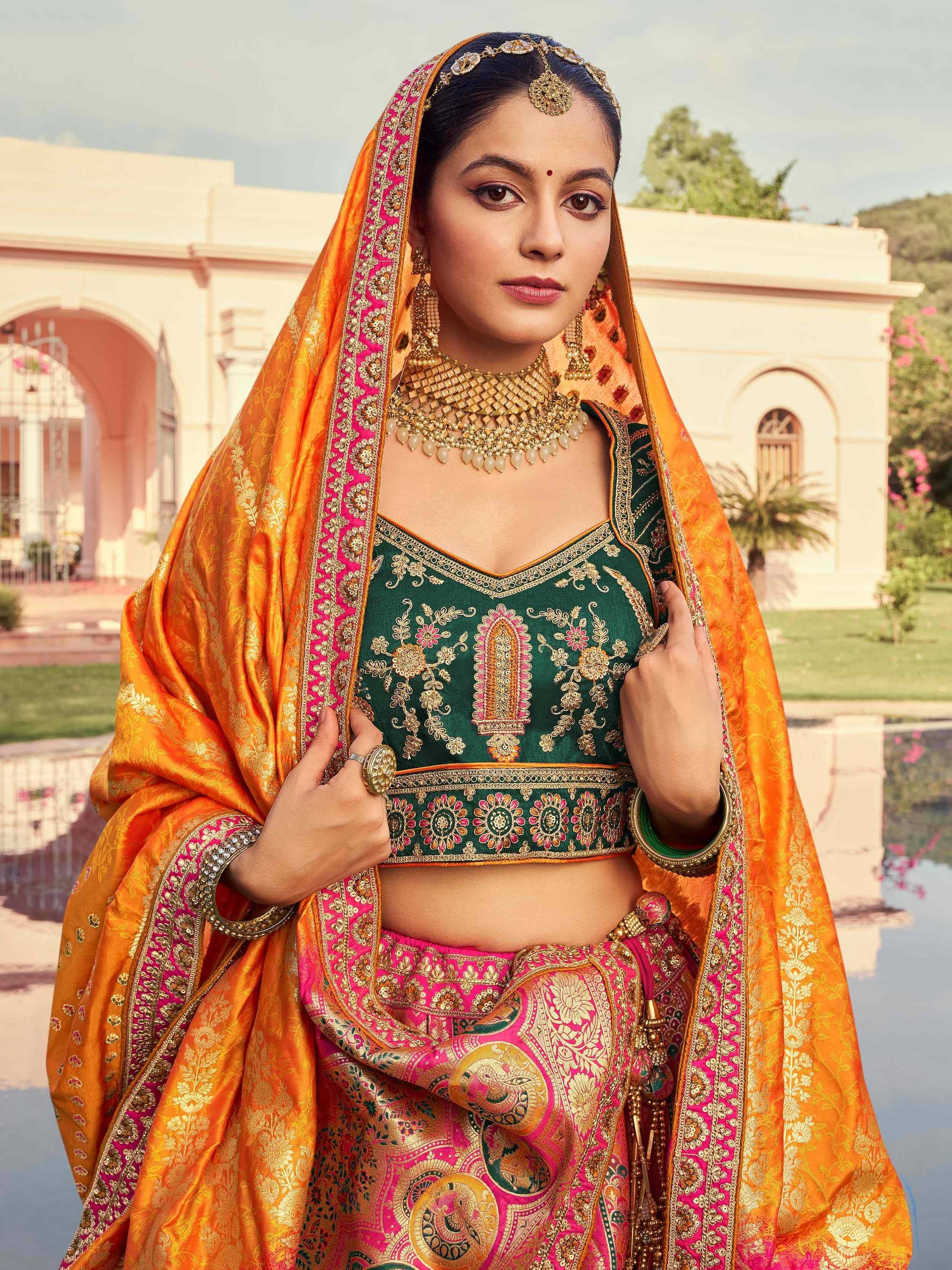 Elegant Indian bridal lehenga choli with green blouse, pink skirt, and orange dupatta, adorned with gold embroidery and traditional jewelry.