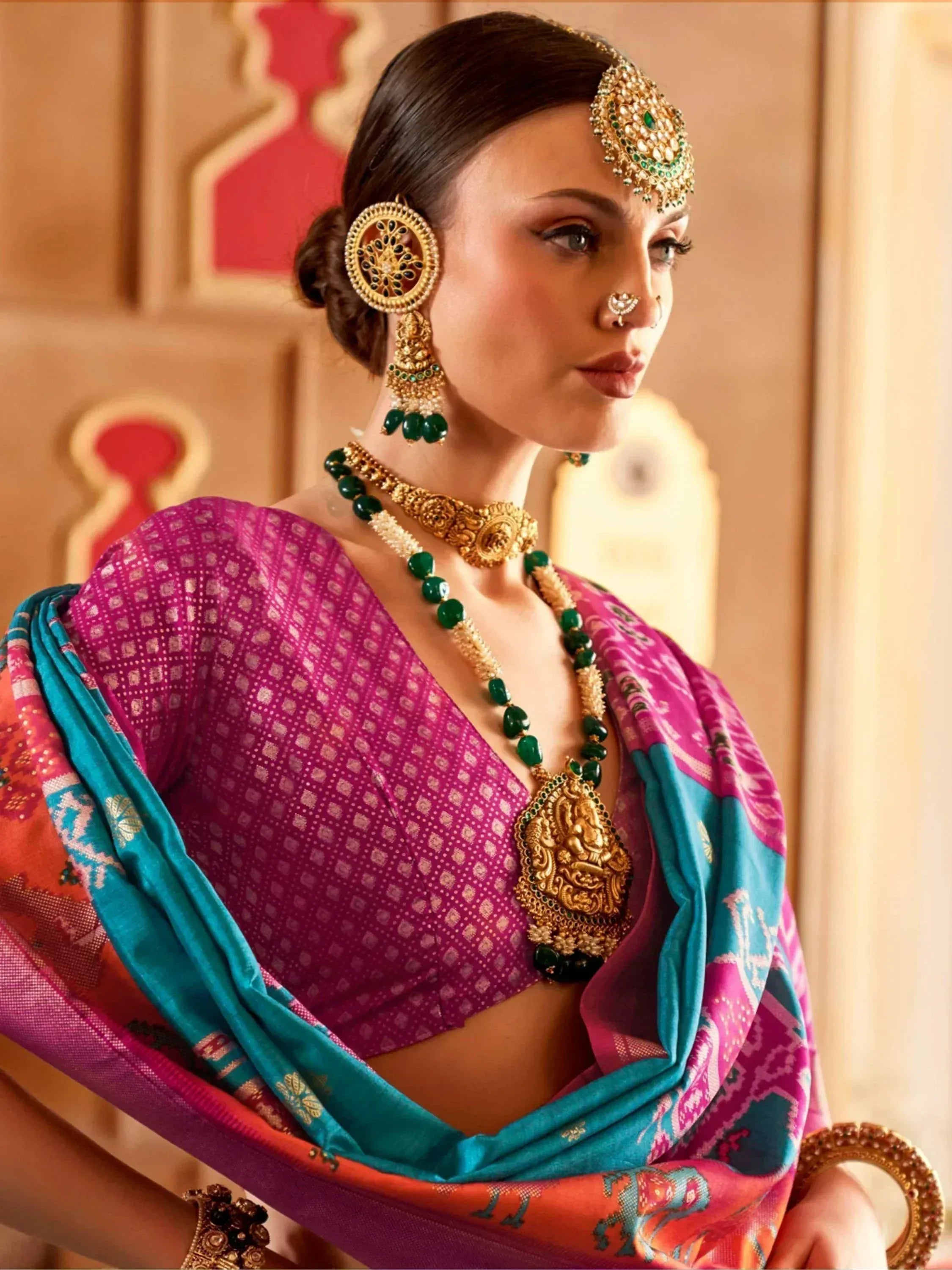 Woman in vibrant pink and teal ethnic saree with gold and green jewelry, traditional Indian attire