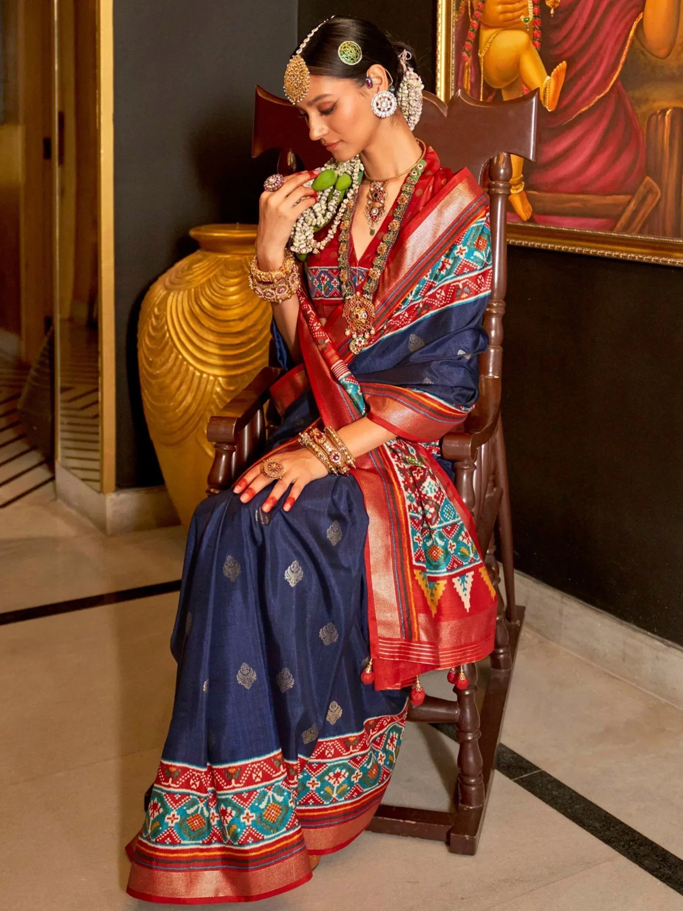 Woman in traditional Indian saree with red and blue embroidery, adorned with ethnic jewelry, seated gracefully.