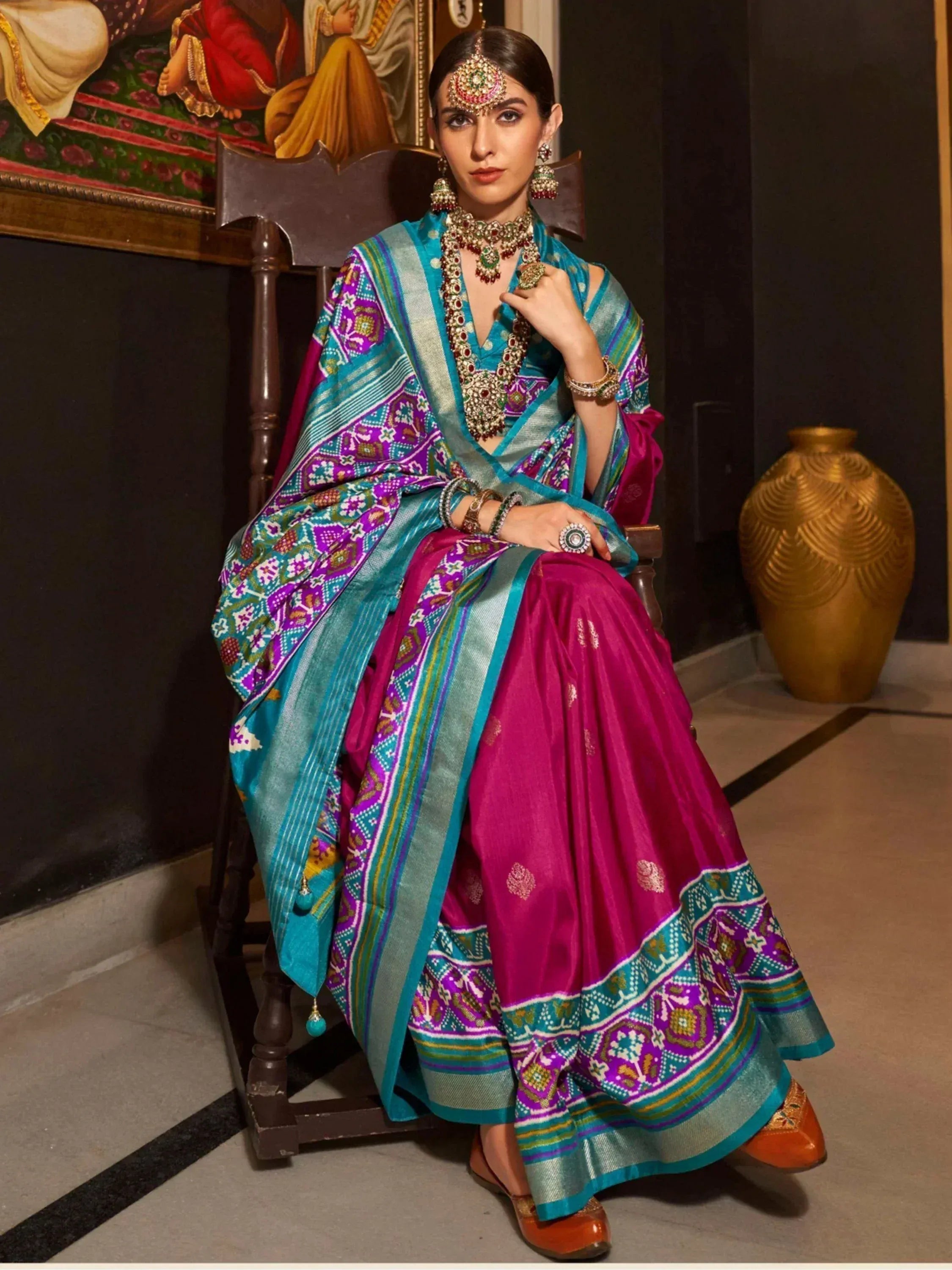 Woman in vibrant Indian ethnic saree with purple and teal patterns, traditional jewelry, seated on wooden chair