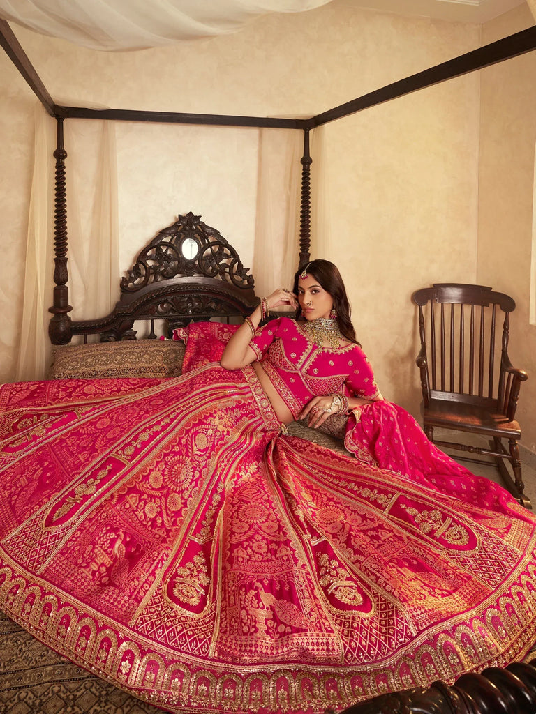 Woman in vibrant pink designer lehenga choli with gold embroidery, seated on ornate bed