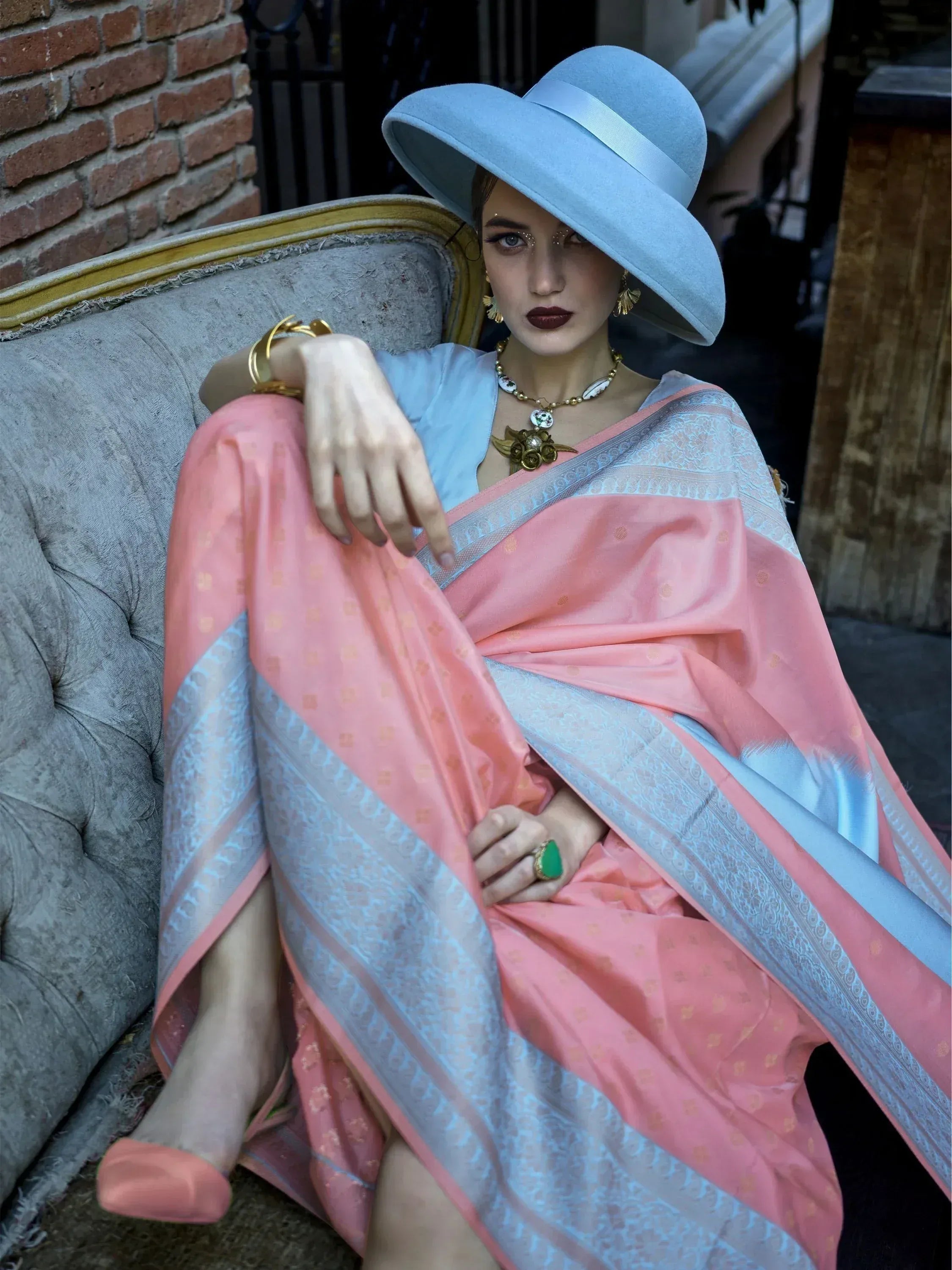 Woman in pink and blue designer saree with wide-brim hat, gold jewelry, sitting on vintage sofa